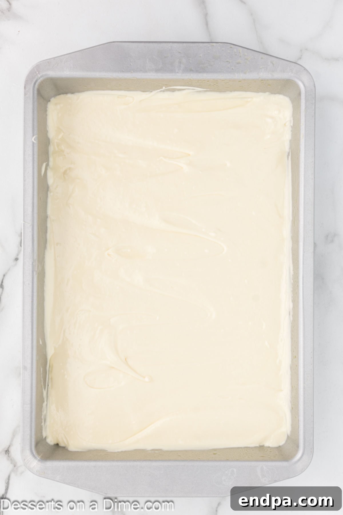 The creamy, white filling batter being spread evenly over the yellow cake crust in the baking dish, ready for baking.