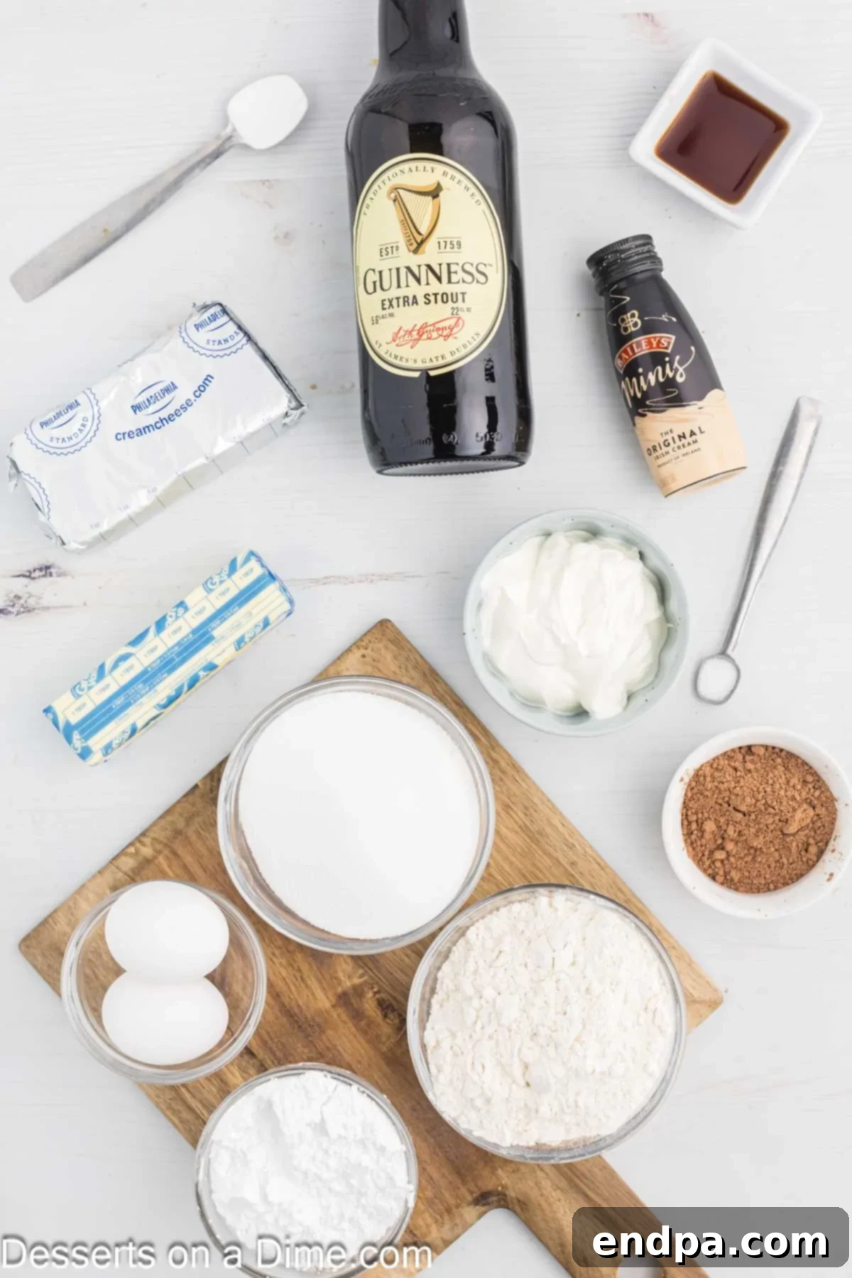 Ingredients for Guinness Cupcakes laid out on a white surface, including flour, cocoa powder, baking soda, butter, eggs, vanilla extract, sour cream, Guinness beer, cream cheese, powdered sugar, Bailey's Irish Cream, and chocolate shavings.