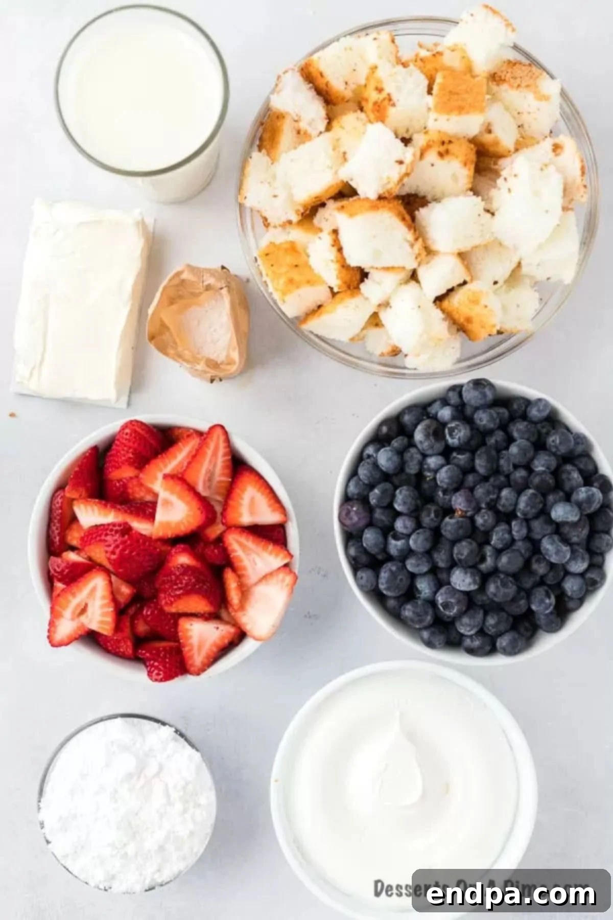 A collection of ingredients laid out, including cubed angel food cake, cream cheese, vanilla pudding mix, and fresh strawberries and blueberries, ready to be assembled into a Red White and Blue Trifle.