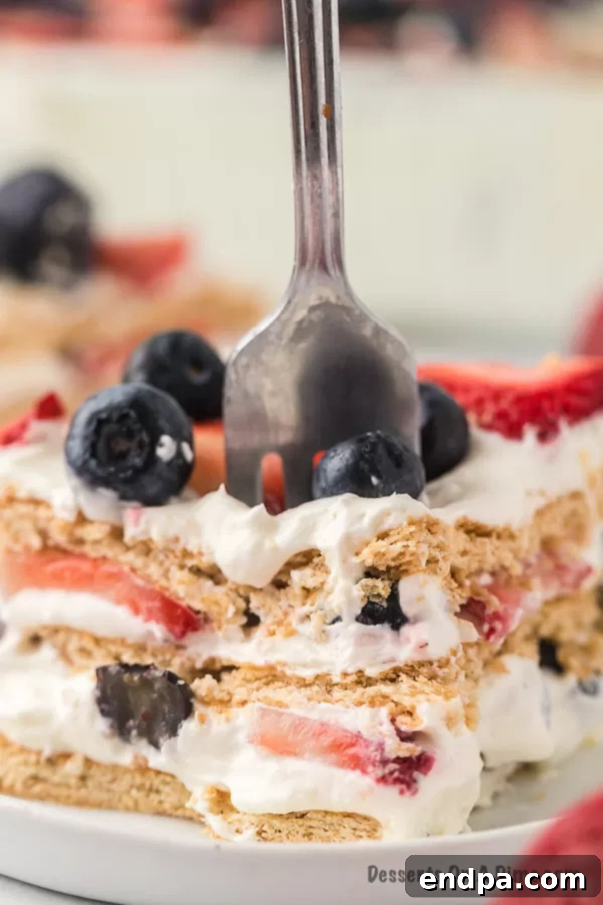 A fork lifts a small piece of a large slice of Red White and Blue Icebox Cake, revealing distinct layers of cream, graham cracker, and berries.