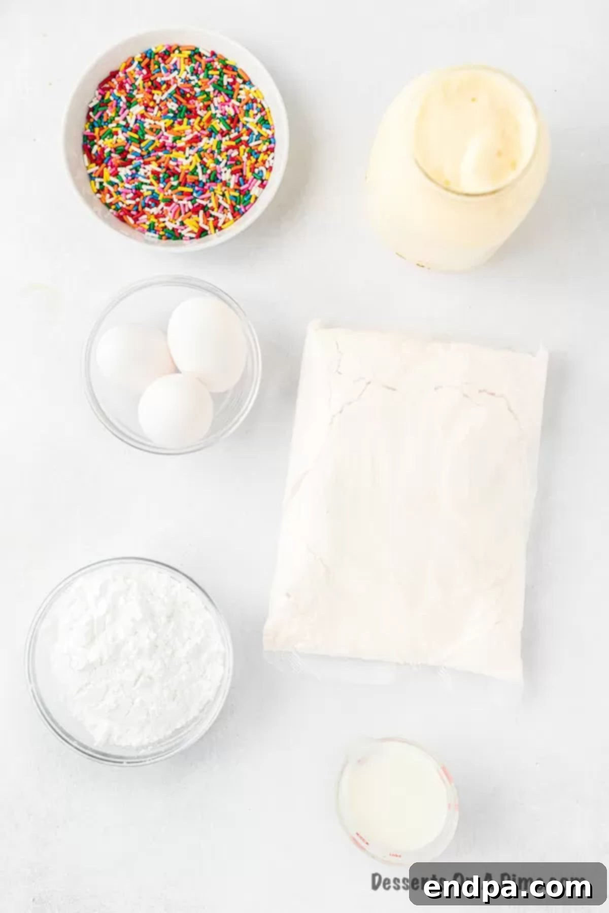 Velvet Ice Cream Cake 3 A flat lay photograph showcasing the simple ingredients for melted ice cream cake: a box of white cake mix, a bowl of melted ice cream, fresh eggs, powdered sugar, a glass of milk, and a colorful container of sprinkles, all ready for baking.