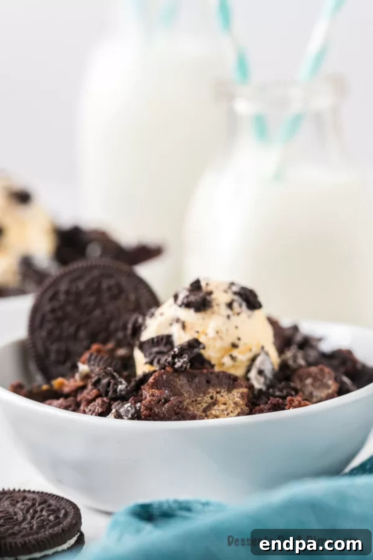 Irresistible Oreo Dump Cake 11 A warm, inviting serving of Oreo Dump Cake in a white bowl, exquisitely topped with melted vanilla ice cream and crunchy crushed Oreos, with a refreshing glass of milk elegantly placed in the background, completing a perfect dessert scene.