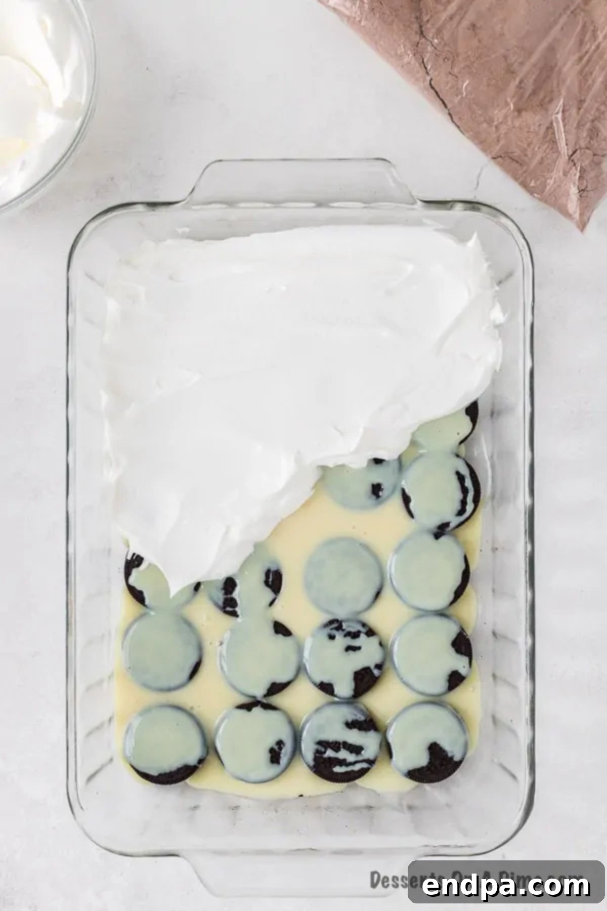 Irresistible Oreo Dump Cake 7 A creamy layer of thawed whipped topping (Cool Whip) being smoothly and evenly spread over the sweetened condensed milk and Oreo base in the baking pan, creating a soft, inviting middle layer.
