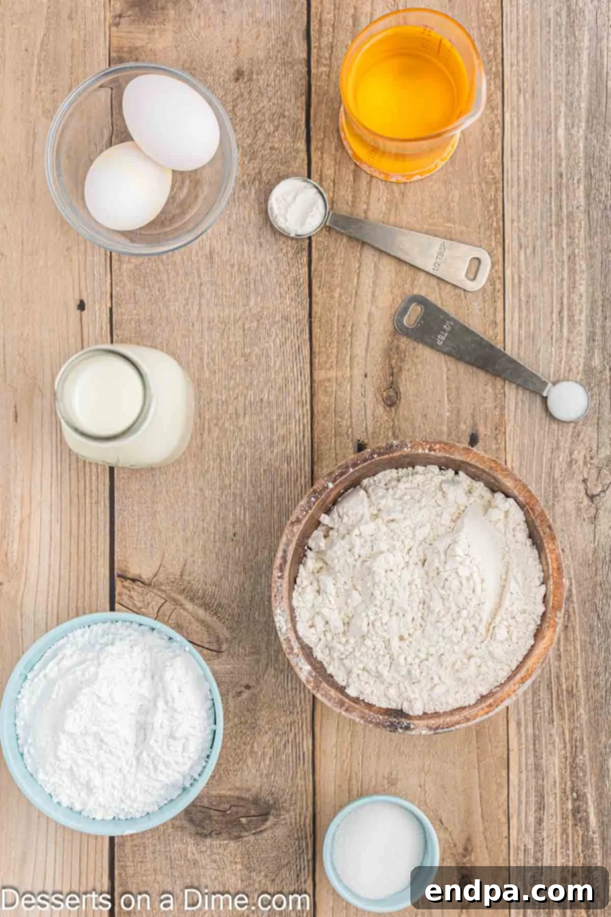 A flat lay of ingredients laid out for making funnel cake bites, including a bowl of all-purpose flour, white sugar, baking powder, a salt shaker, a jug of milk, two eggs in their shells, a bottle of vegetable oil, and a bowl of powdered sugar.