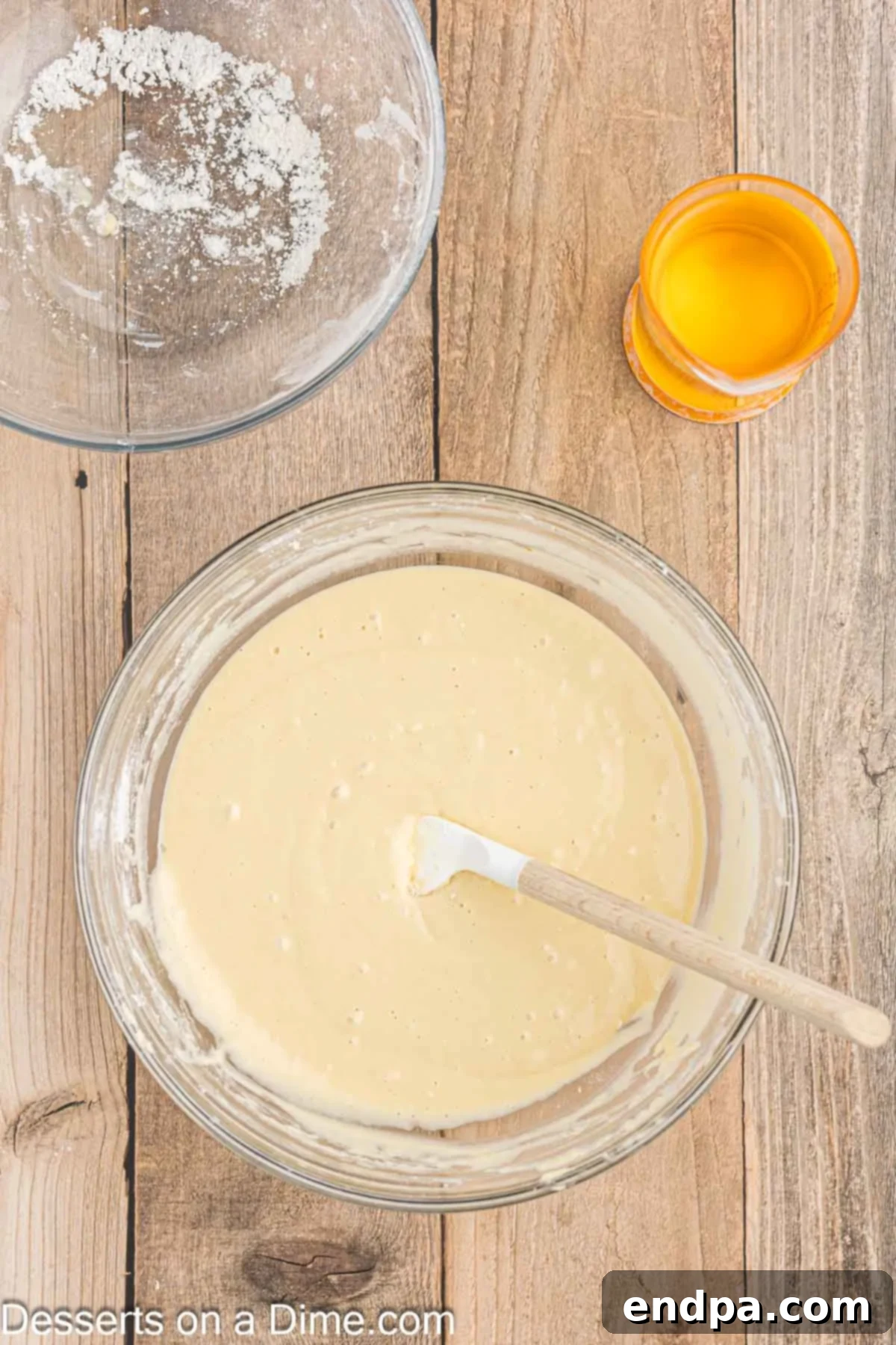 A clear glass bowl showing wet ingredients (milk and eggs) mixed with dry ingredients, forming a smooth, pale yellow funnel cake batter.