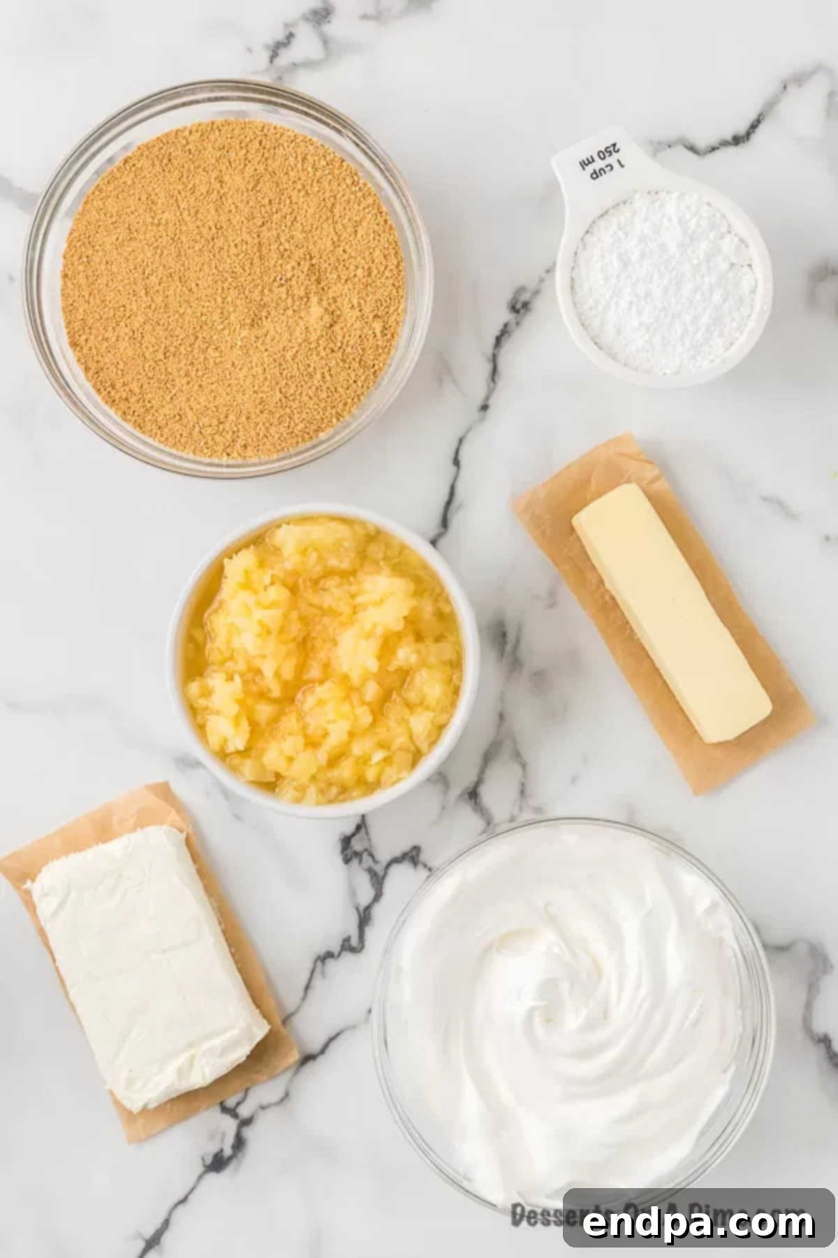 A flat lay photograph showcasing the ingredients for Pineapple Dream Dessert: graham cracker crumbs, powdered sugar, sticks of butter, blocks of cream cheese, a can of crushed pineapple, and a tub of whipped topping.