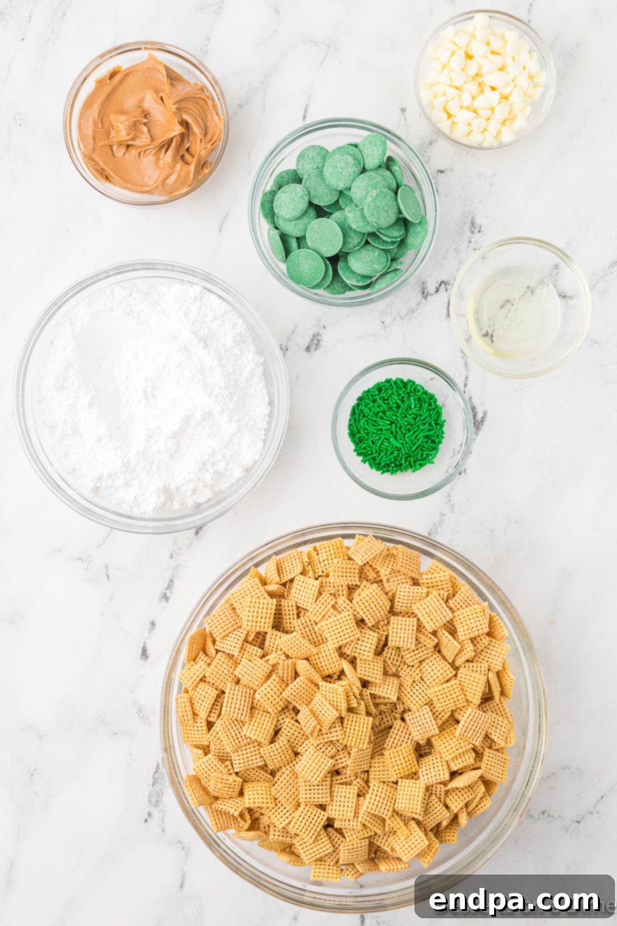 A collection of ingredients laid out for St. Patrick's Day Puppy Chow, including a box of Chex cereal, a jar of peanut butter, green candy melts, a bowl of powdered sugar, green sprinkles, and green M&M's candies.