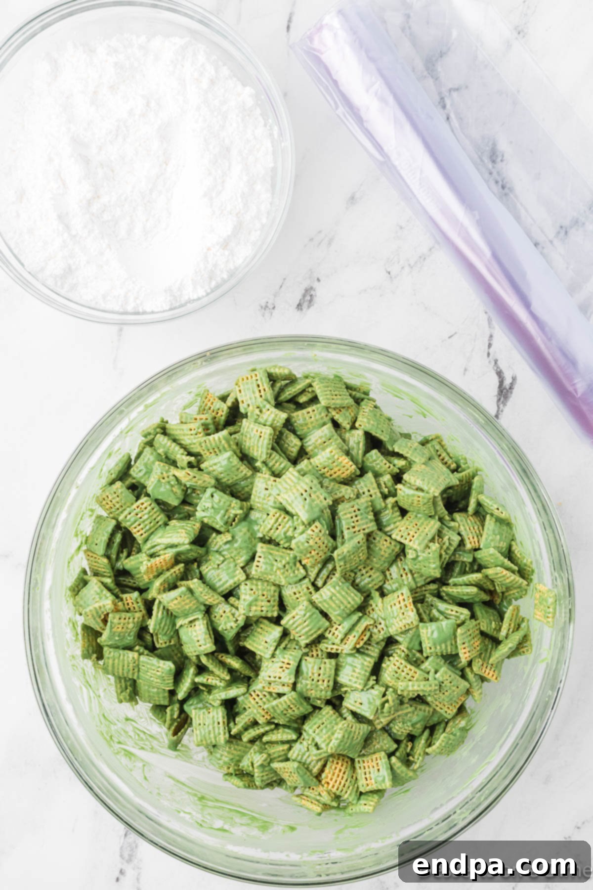 Large mixing bowl with Chex cereal being coated with the melted green peanut butter and candy mixture, showing an even distribution.