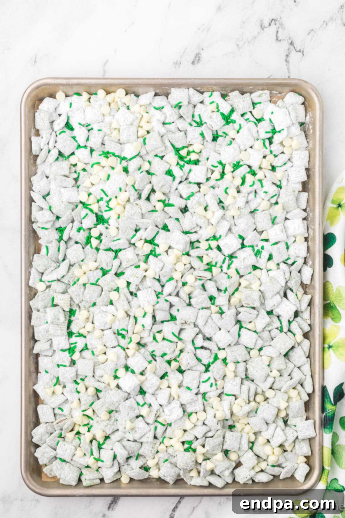 Finished St. Patrick's Day Puppy Chow spread on parchment paper, dusted with powdered sugar, and topped with white chocolate chips, green M&M's, and festive green sprinkles.