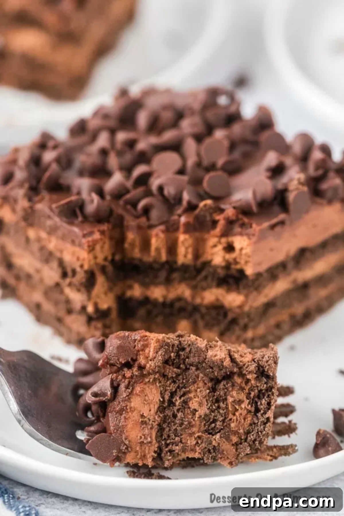 A slice of chocolate icebox cake with ganache and mini chocolate chips, served on a white plate with a fork.