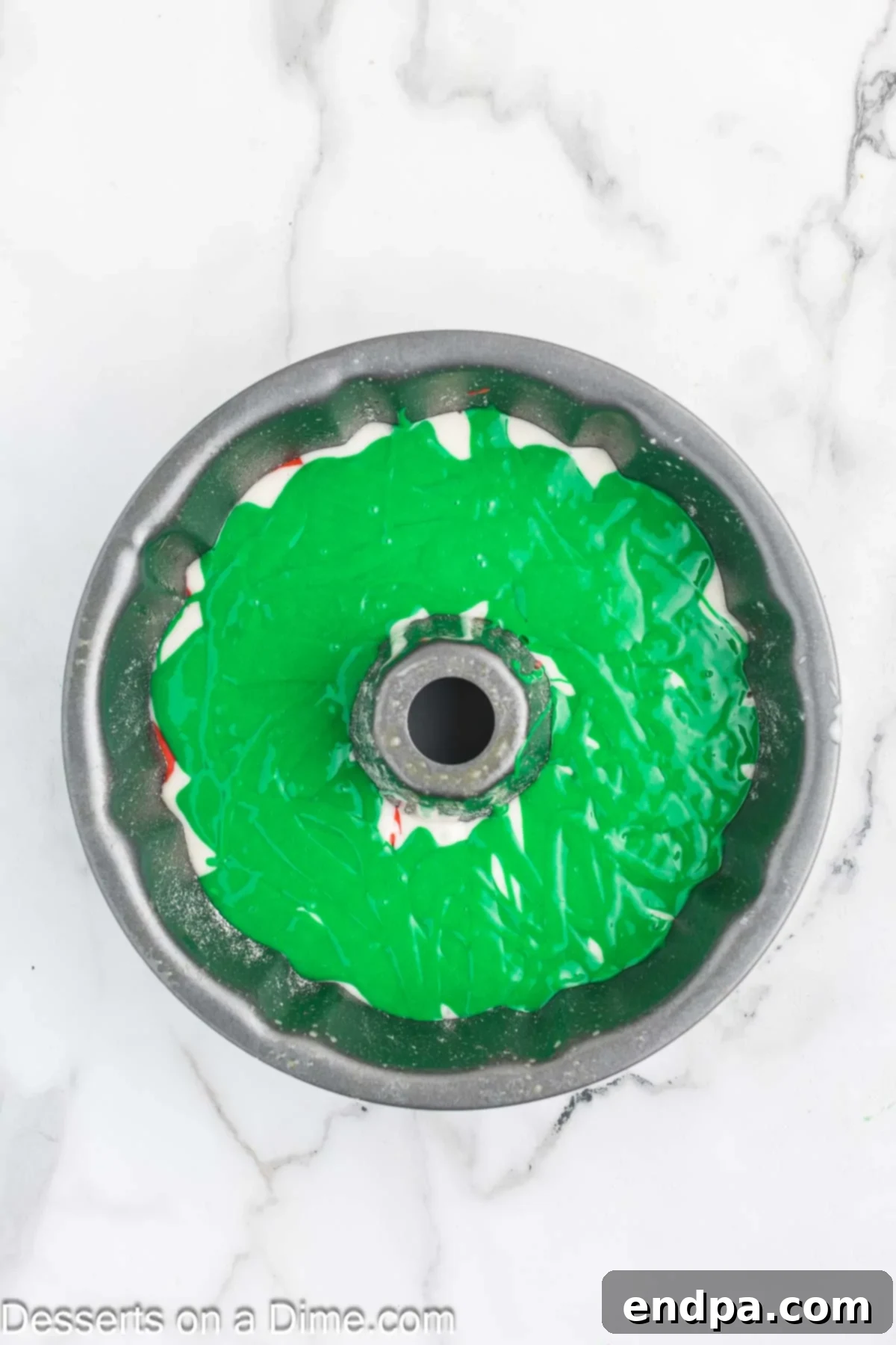 Green cake batter dolloped on top of the red batter in the Bundt pan, ready for swirling.