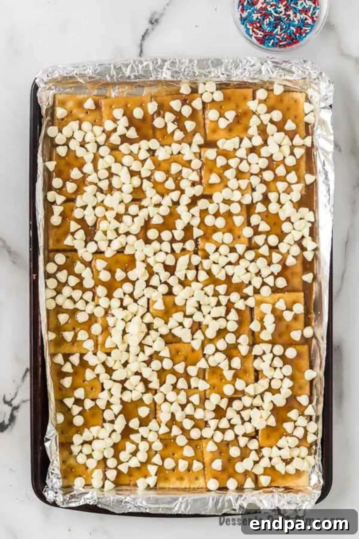 A generous layer of white chocolate chips sprinkled directly onto the hot, bubbly cracker toffee, poised to melt into a smooth, creamy topping for the patriotic bark.