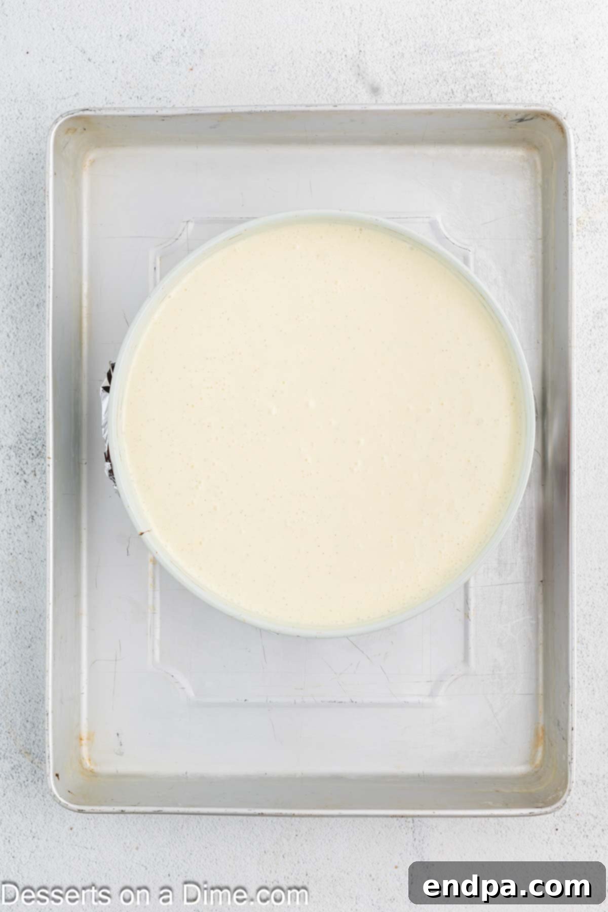 Crispy Caramel Cheesecake 9 Cheesecake batter poured over the crust in a springform pan, wrapped in aluminum foil, and placed in a water bath inside a larger roasting pan, ready for baking.