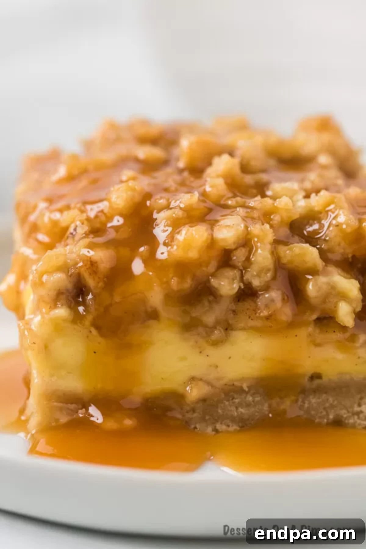 Close-up image of a decadent Caramel Apple Cheesecake bar with a glistening caramel drizzle on a pristine white plate, showcasing its luscious layers.