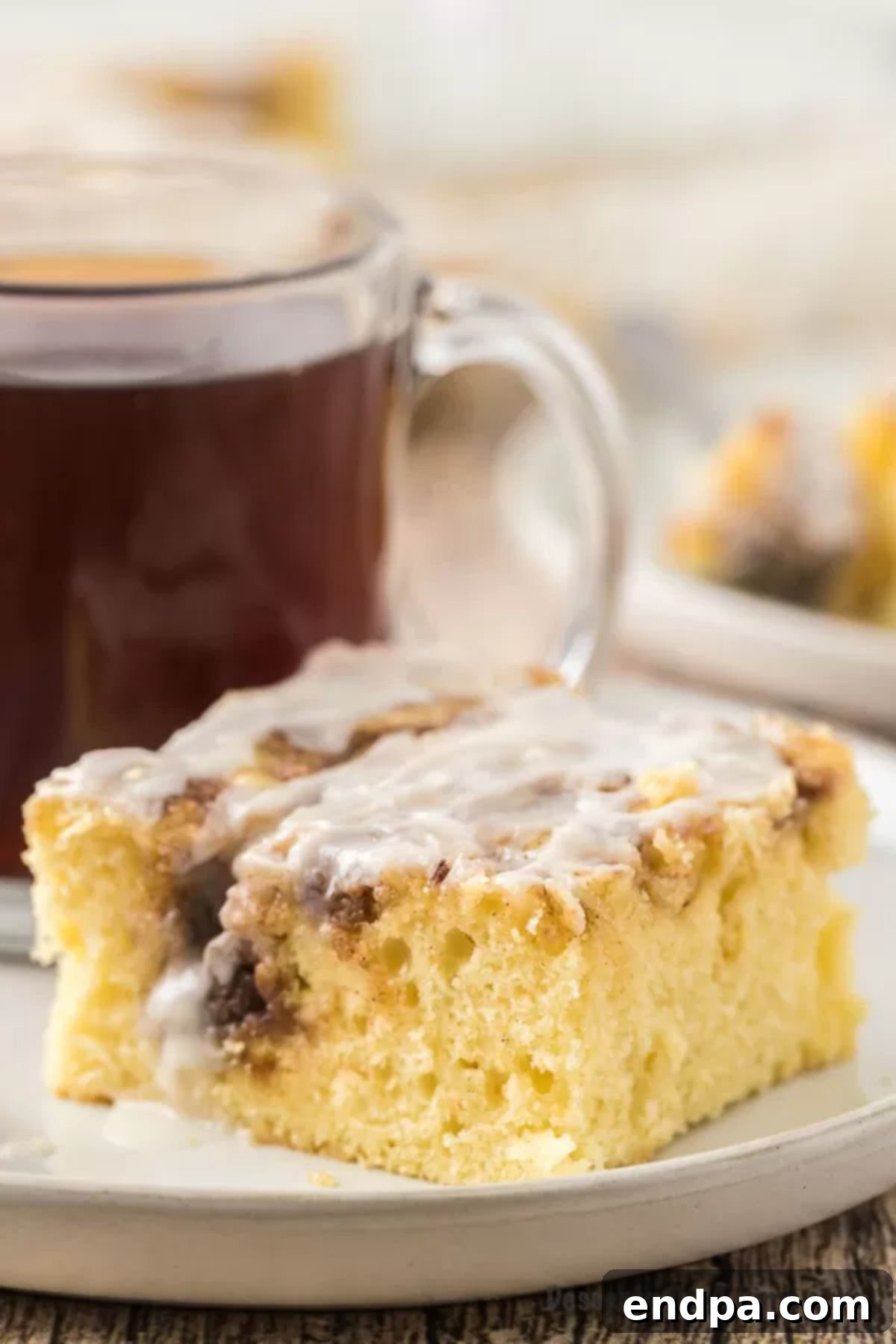 Indulgent Cinnamon Roll Cake Delight 2 A piece of the cinnamon roll cake on a white plate with a cup of coffee behind it
