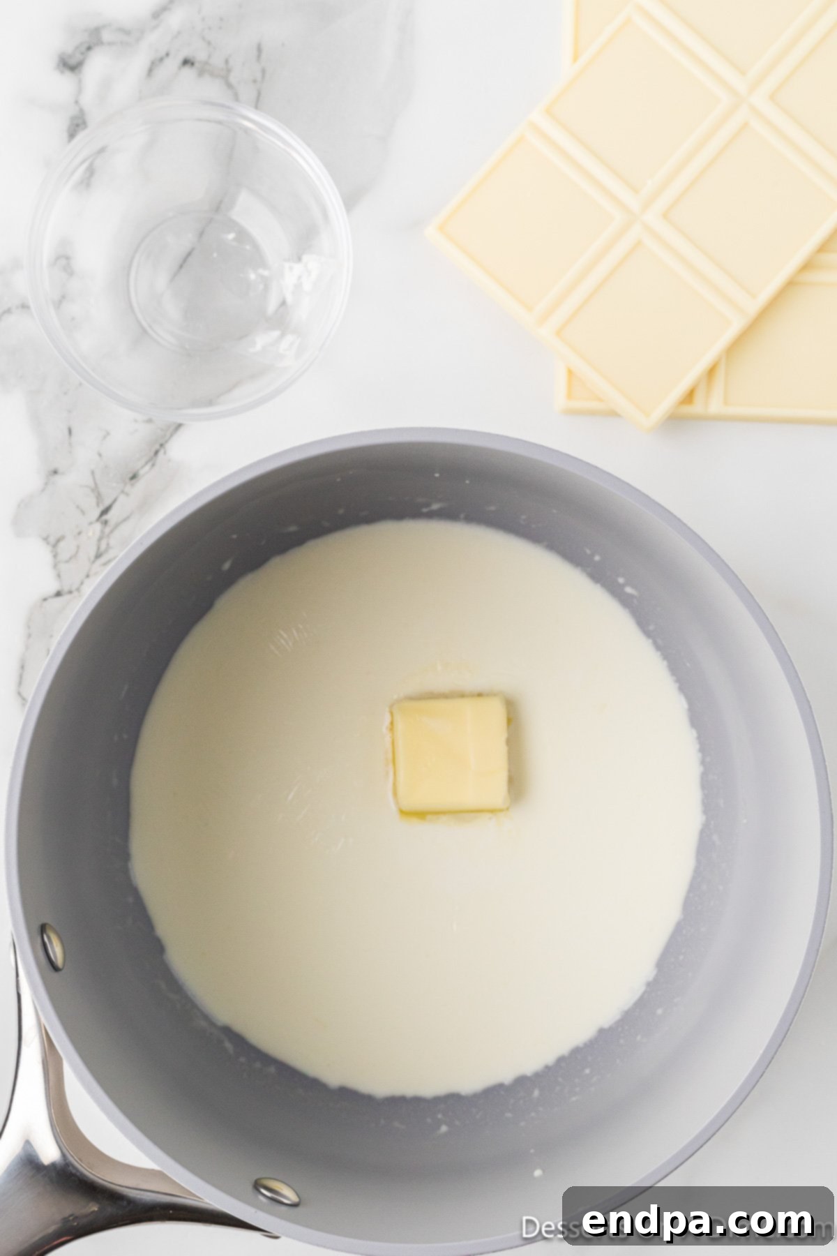 A tablespoon of butter being added to the simmering heavy cream in a saucepan.