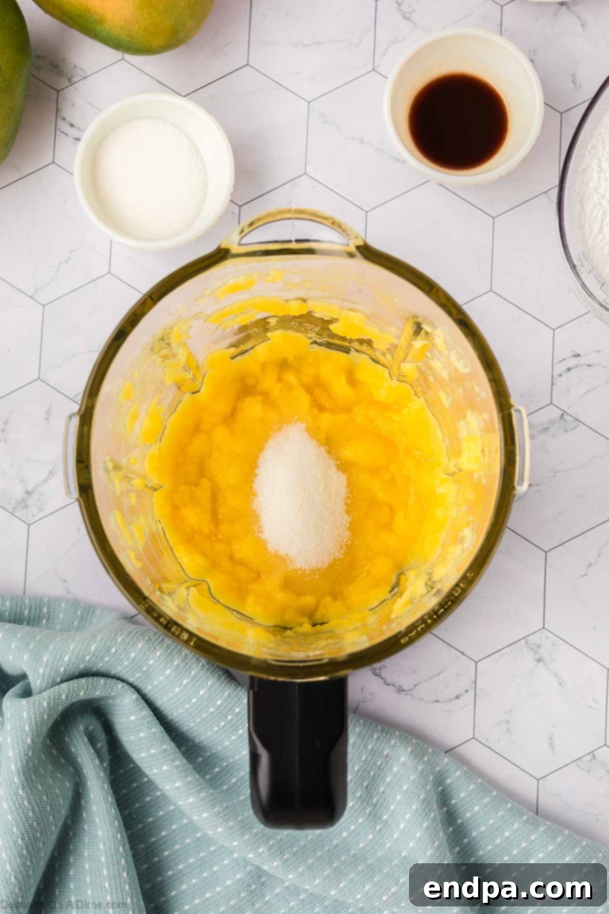 Fresh mango chunks and granulated sugar being pureed in a blender or food processor until smooth for the cake filling.