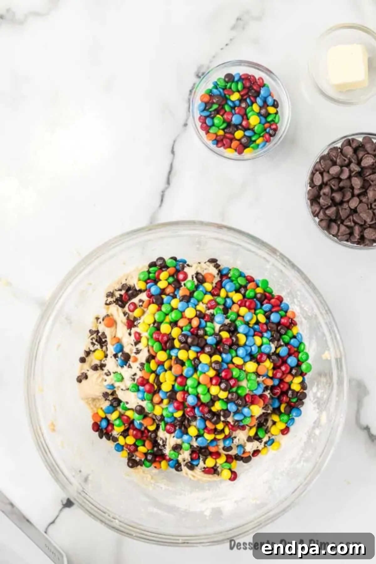 Miniature M&M's and chocolate chips being added to the cookie dough mixture in a bowl.