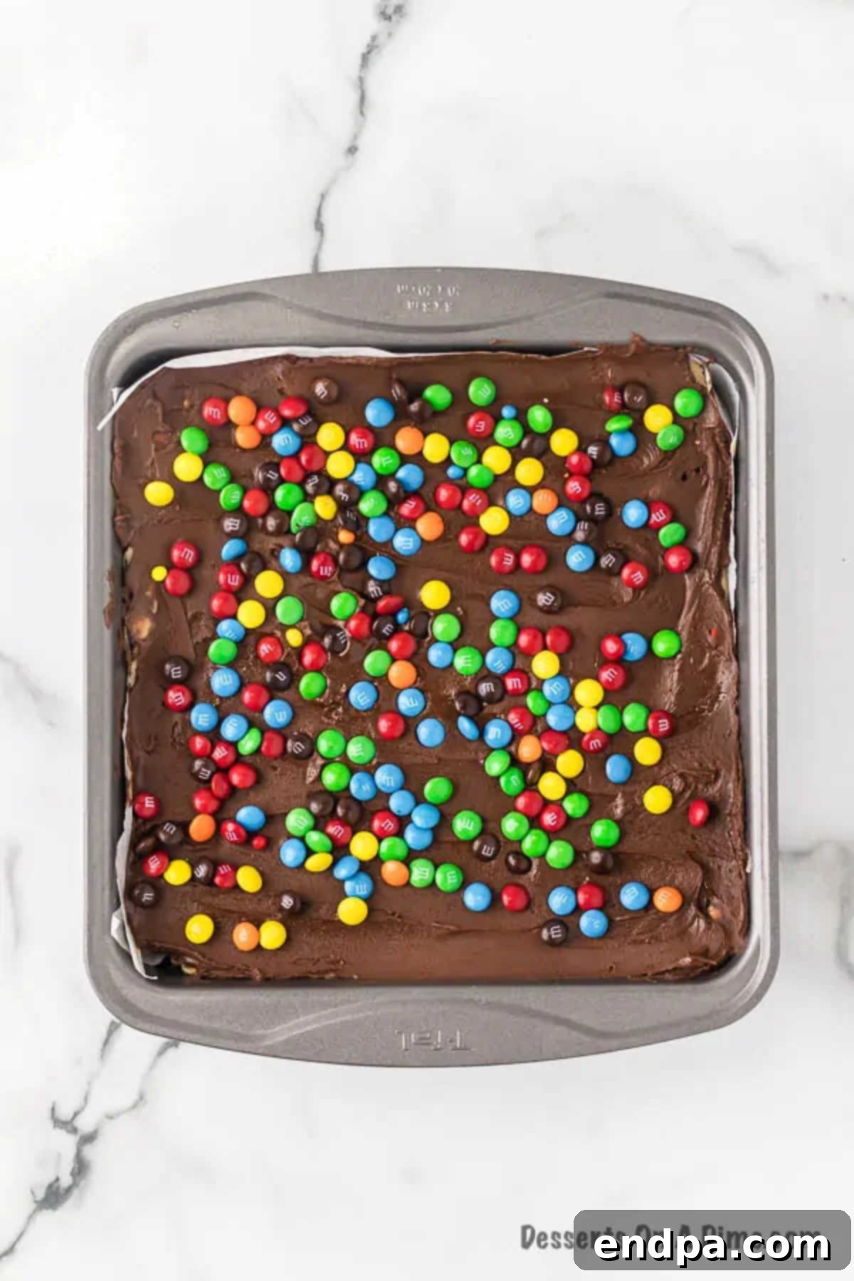 Melted chocolate topping being spread evenly over the cookie dough base and sprinkled with M&M's.