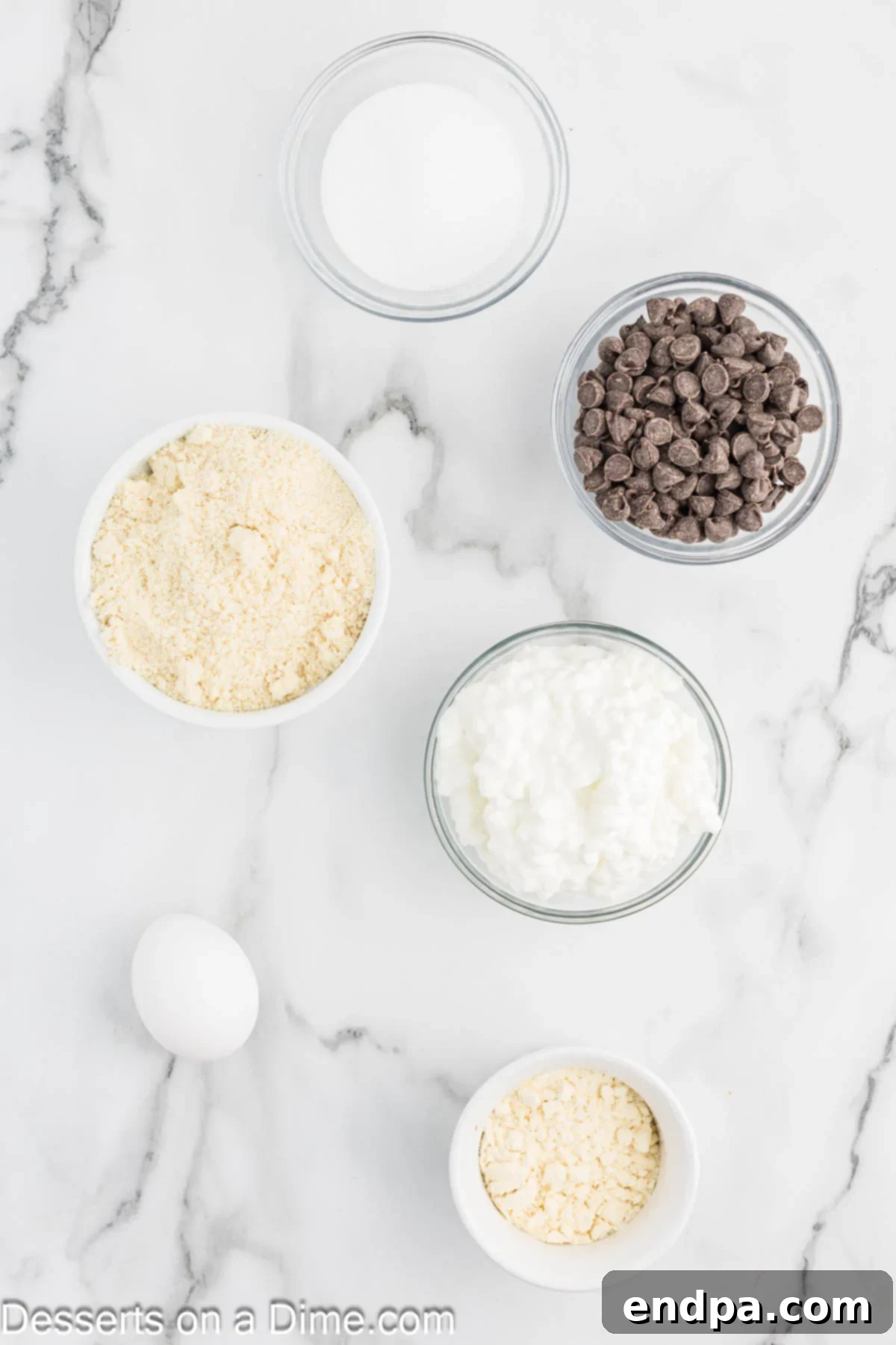 Individual ingredients for cottage cheese cookies laid out: a bowl of whole fat cottage cheese, a large egg, monk fruit sweetener, almond flour, vanilla protein powder, and sugar-free chocolate chips.