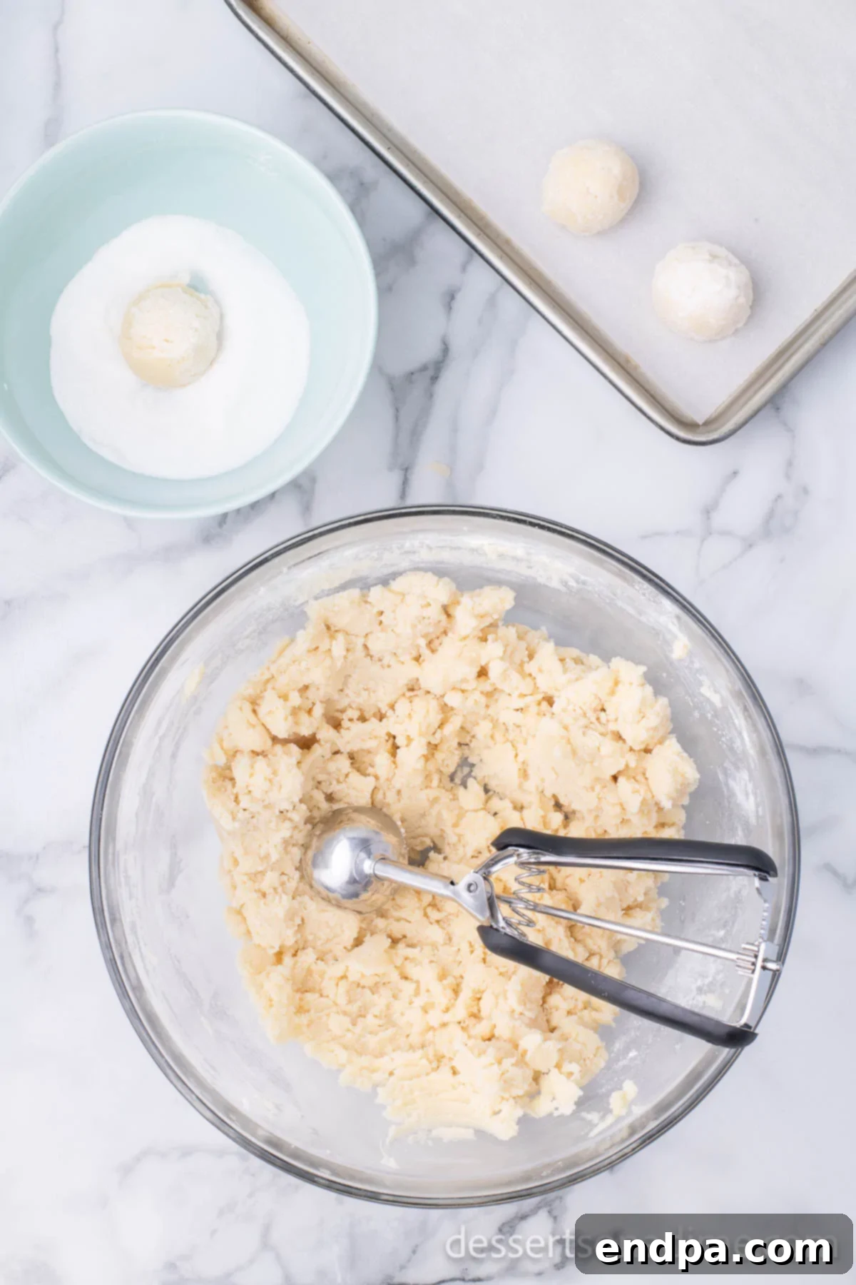THUMBPRINT JAM COOKIES 8 Cookie dough balls being rolled in granulated sugar on a baking sheet, with a cookie scoop nearby.