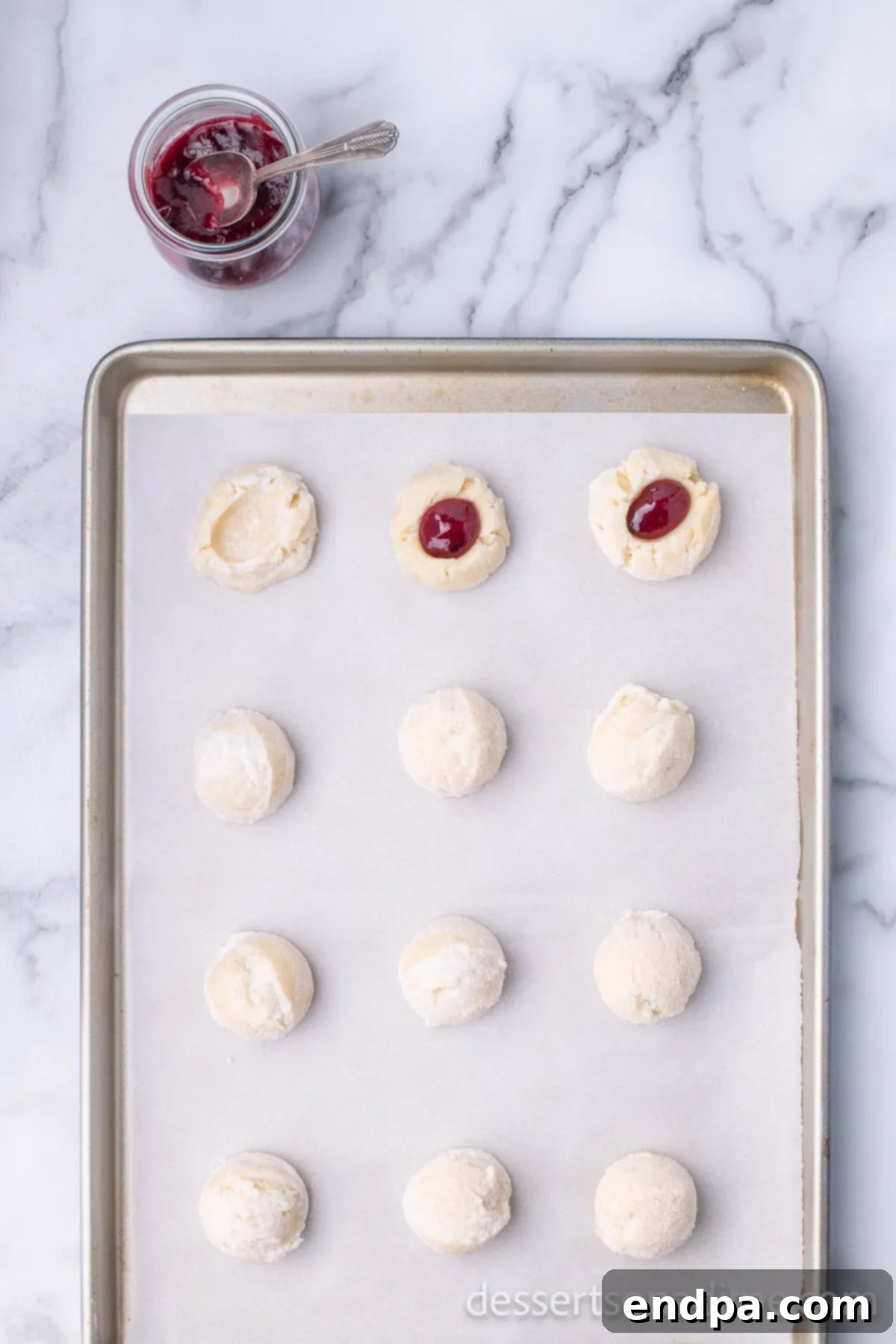 THUMBPRINT JAM COOKIES 9 Baking sheet with shaped cookie dough balls, each with a thumbprint indentation.