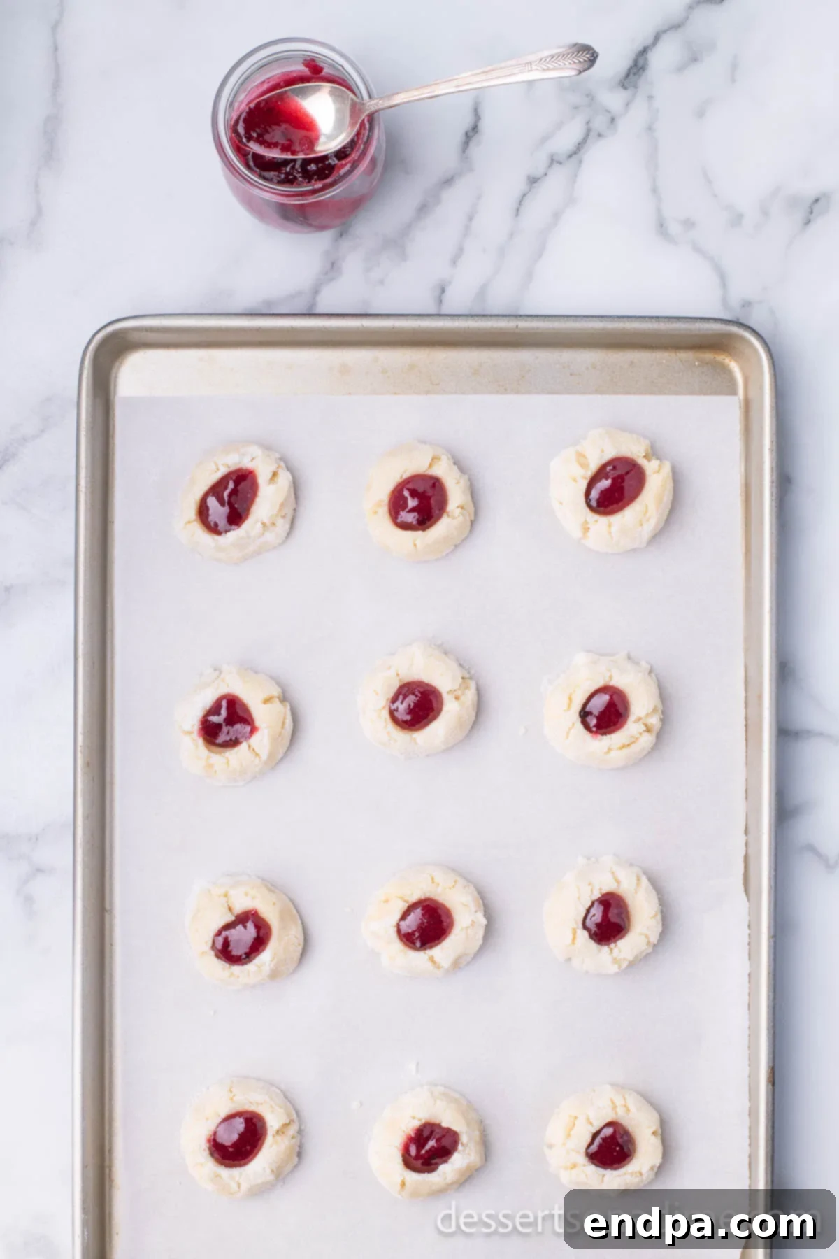 THUMBPRINT JAM COOKIES 10 Baking sheet with thumbprint cookies, each indentation filled with vibrant red raspberry jam.