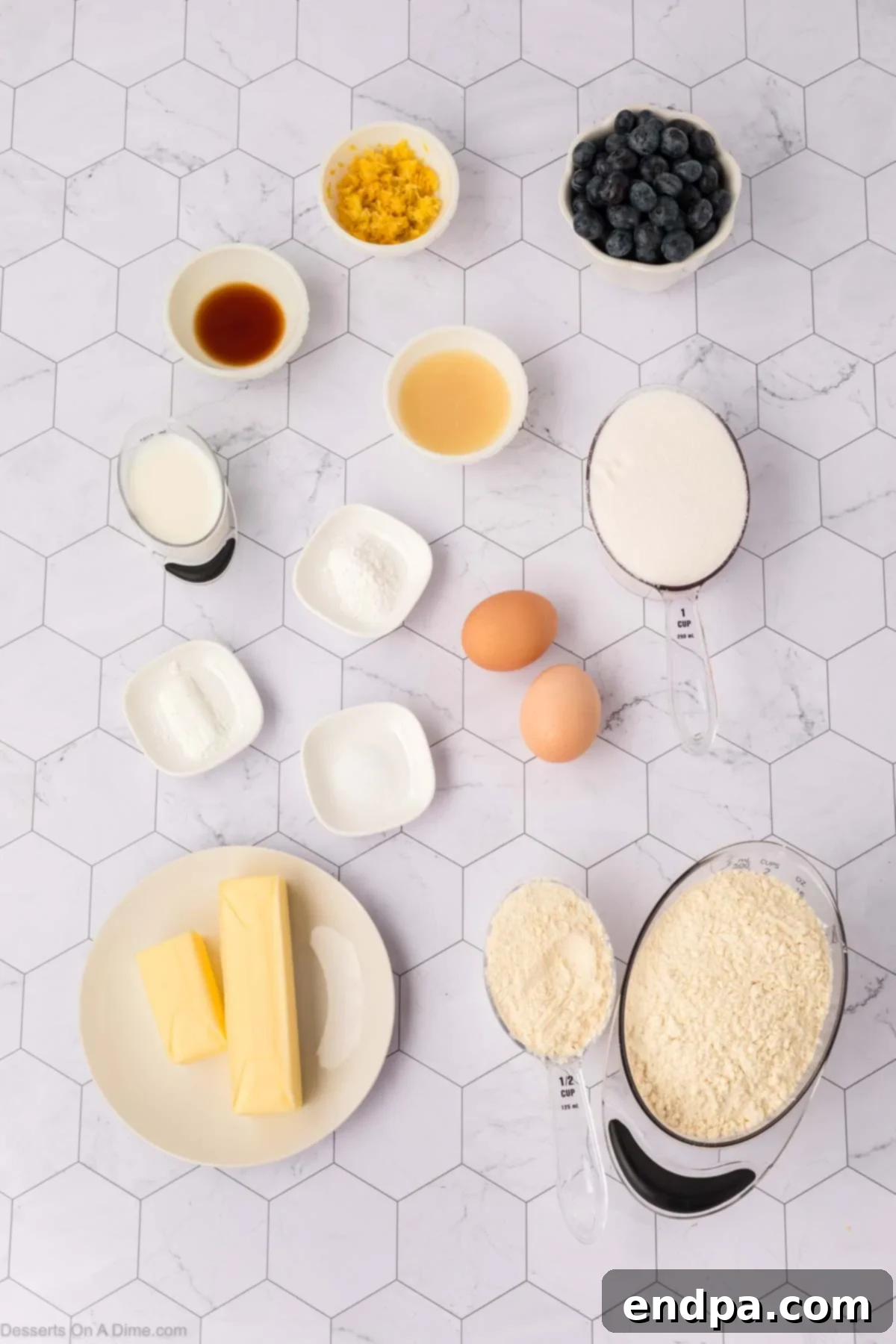 A flat lay showing essential ingredients for lemon blueberry cookies, including flour, butter, eggs, vanilla, fresh lemons, milk, blueberries, and powdered sugar.