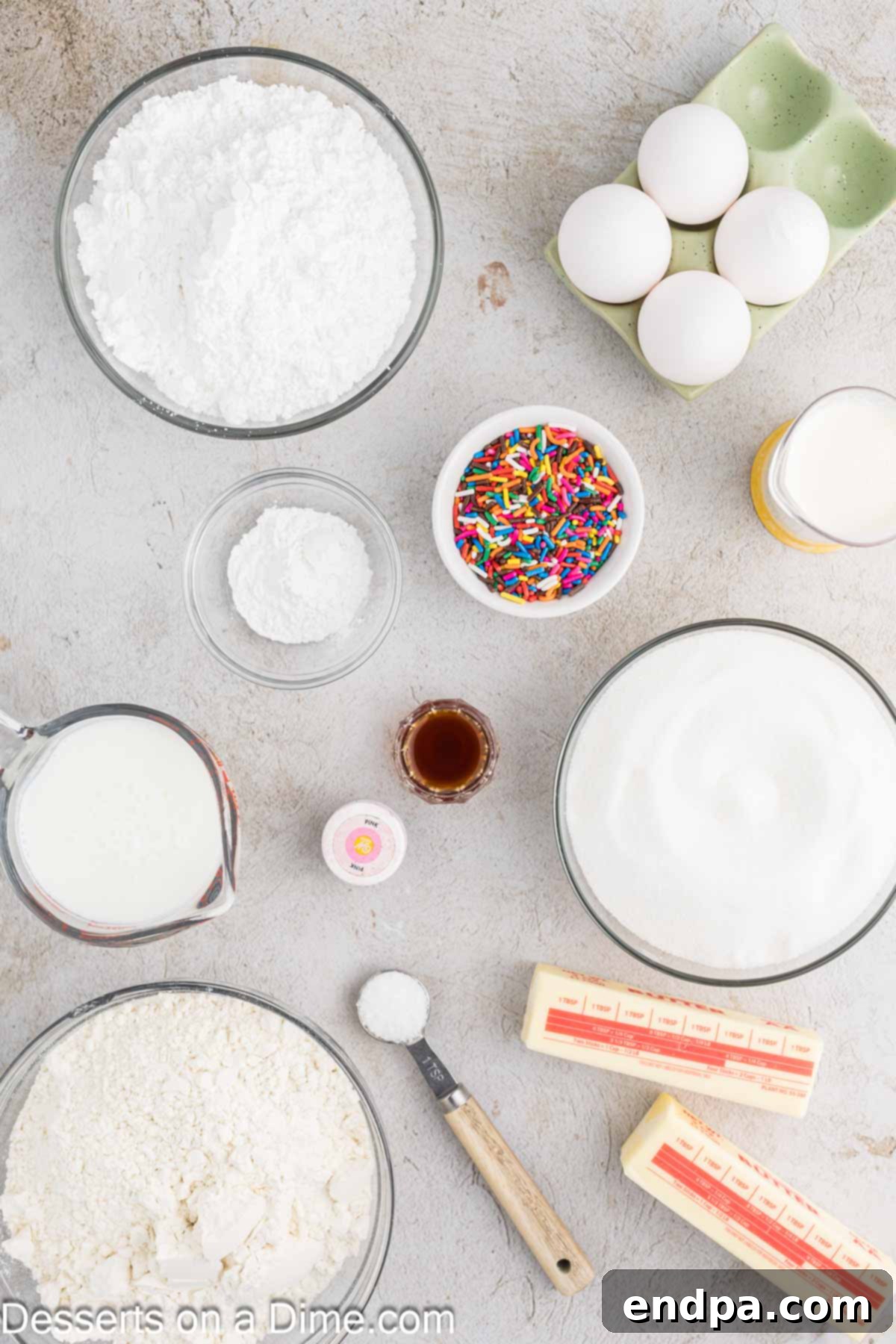 A flat lay of fresh ingredients for baking a donut cake: softened butter, large eggs, vanilla extract, cake flour, baking powder, salt, buttermilk, powdered sugar, heavy cream, food coloring, and a selection of colorful sprinkles.