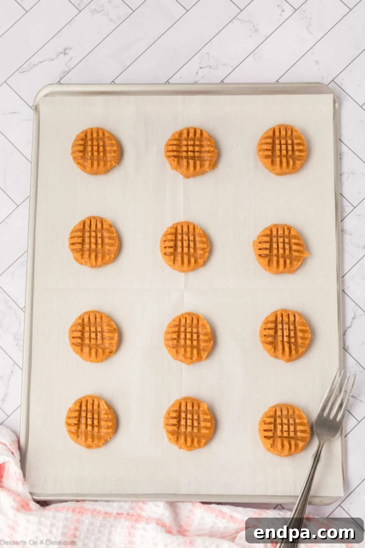 Peanut butter cookies on a baking sheet, each with a classic criss-cross pattern pressed into the top.
