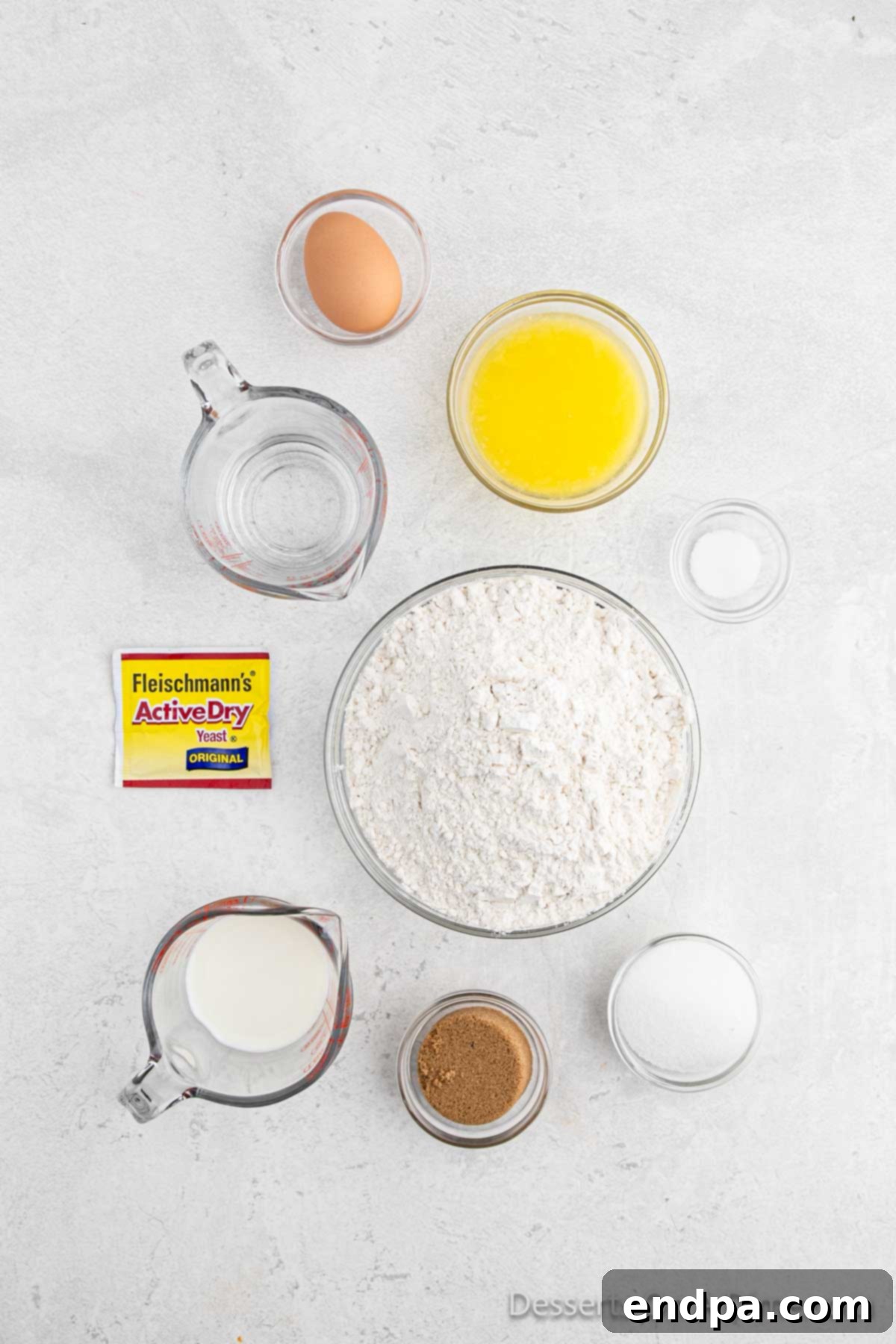 Assortment of key ingredients for cinnamon rolls displayed on a wooden surface: active dry yeast, milk, butter, brown sugar, all-purpose flour, and cinnamon sticks.