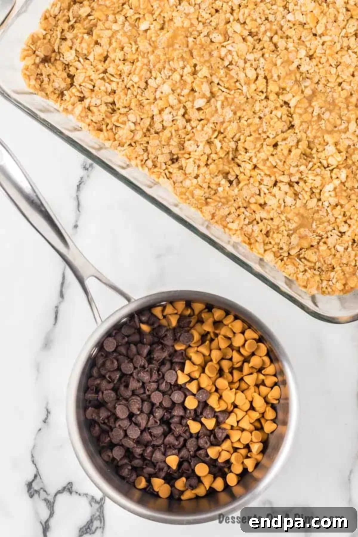 A 9x13 inch baking pan filled with the pressed Rice Krispies and peanut butter base, with a small saucepan next to it containing melting chocolate and butterscotch chips, ready for the topping.