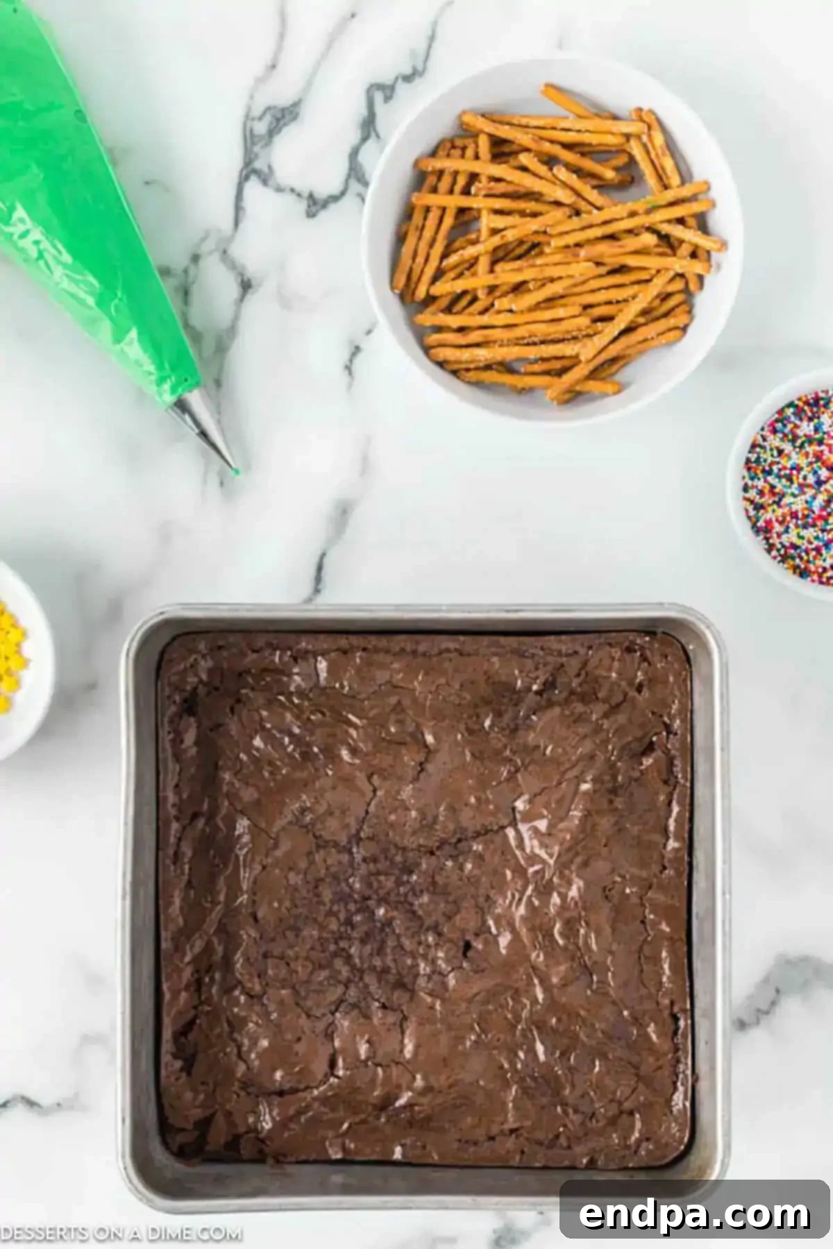 A flat lay image displaying all the simple ingredients required for Christmas Tree Brownies: a box of brownie mix, green icing, various sprinkles, and pretzel sticks.