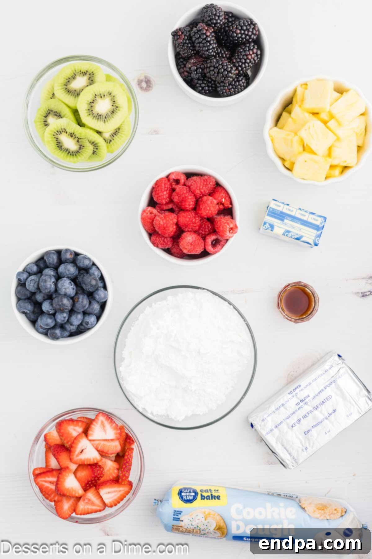 Fresh Fruit Fiesta Pizza 3 All ingredients for a fruit pizza laid out: a roll of sugar cookie dough, a block of cream cheese, butter, vanilla extract, powdered sugar, and a bowl of mixed fresh fruit including strawberries, blueberries, and kiwi.