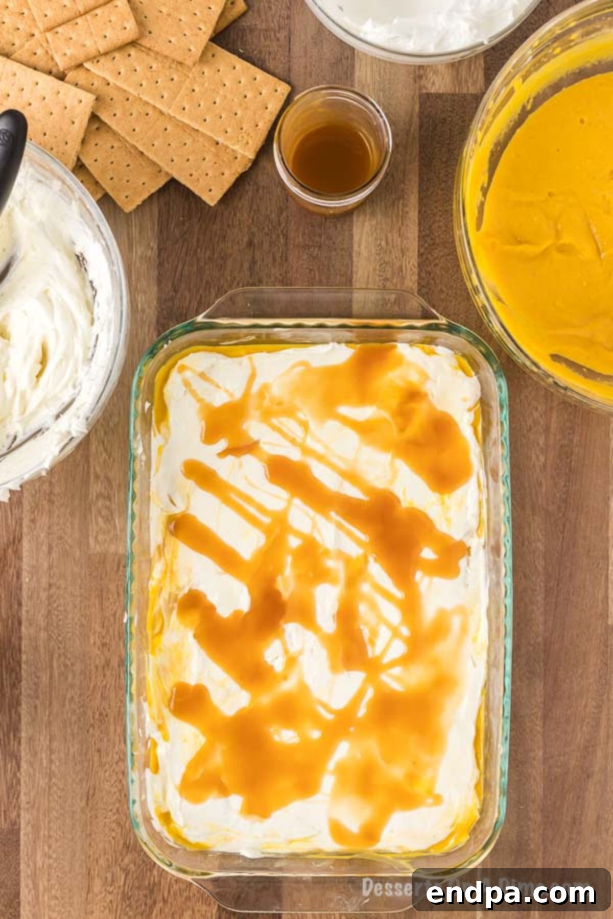 A generous drizzle of golden caramel sauce cascades over the creamy cream cheese layer of the partially assembled pumpkin lasagna in the baking pan, adding a luscious touch before repeating the layers.