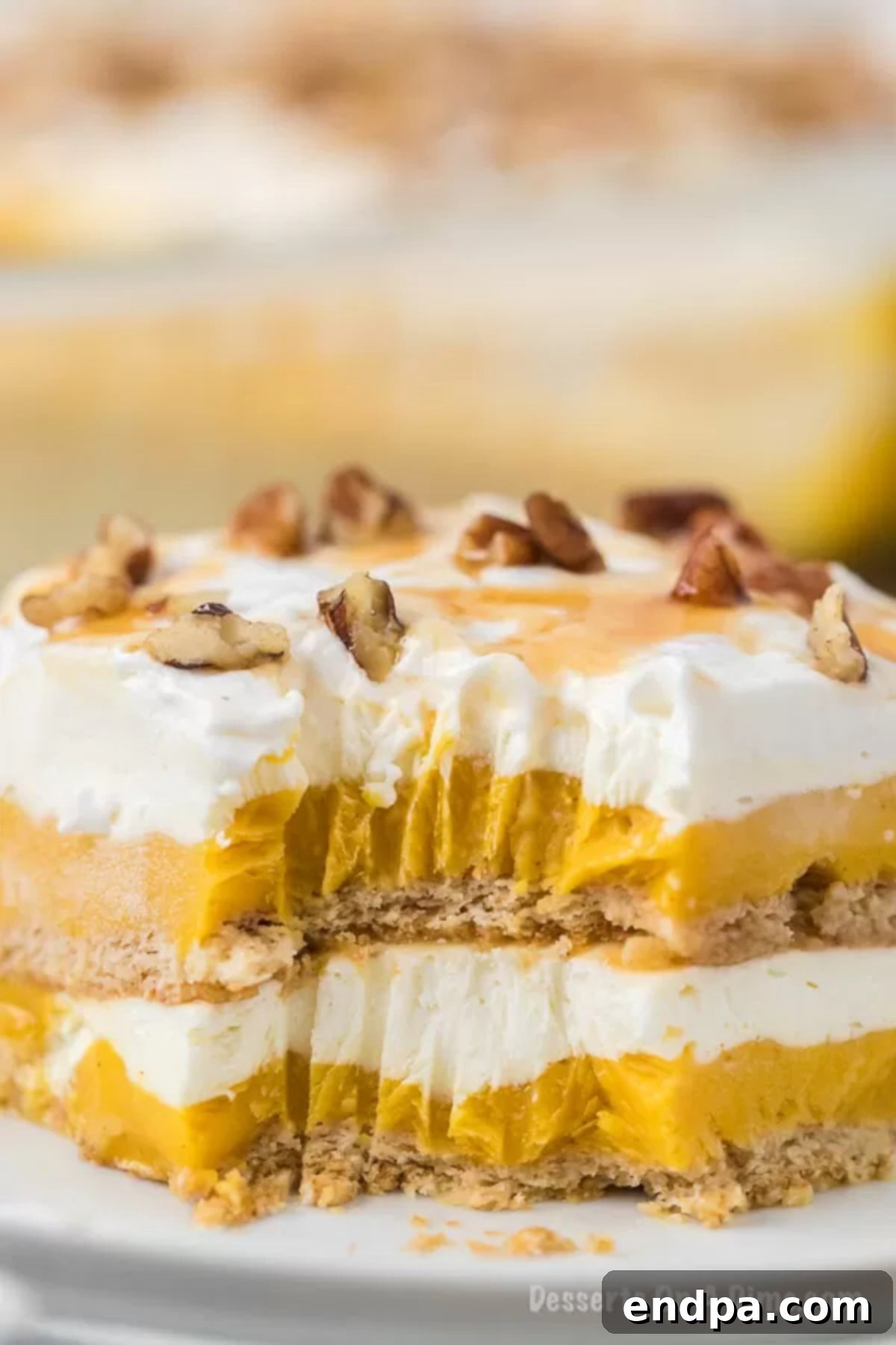A close-up shot of a slice of pumpkin lasagna on a white plate, with a small bite taken from the corner, revealing the creamy layers and crunchy crust. The remaining caramel drizzle and pecans are visible on top.
