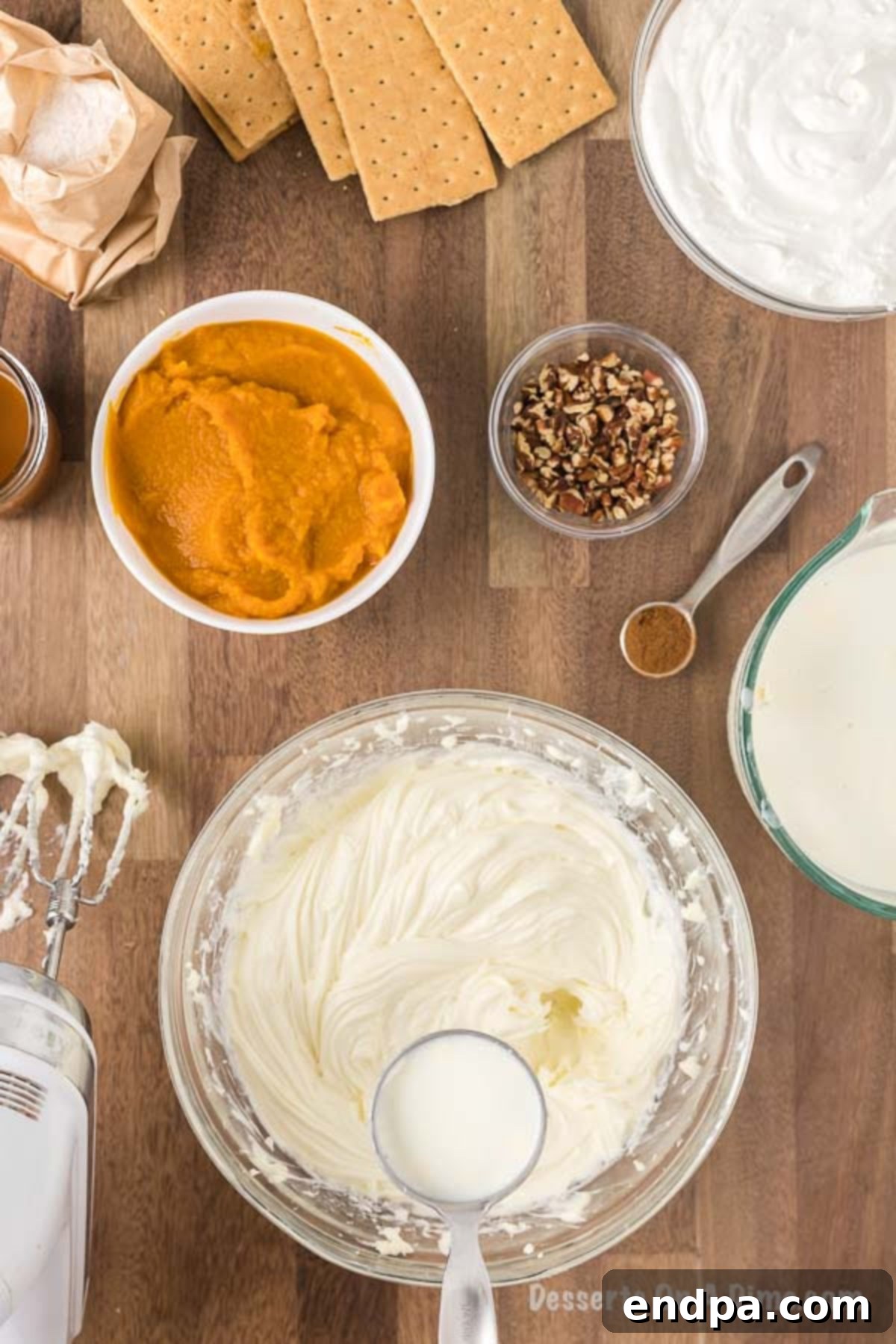 A stream of milk is being slowly poured into the fluffy cream cheese and sugar mixture in a large mixing bowl, as it continues to be beaten to ensure smooth incorporation.