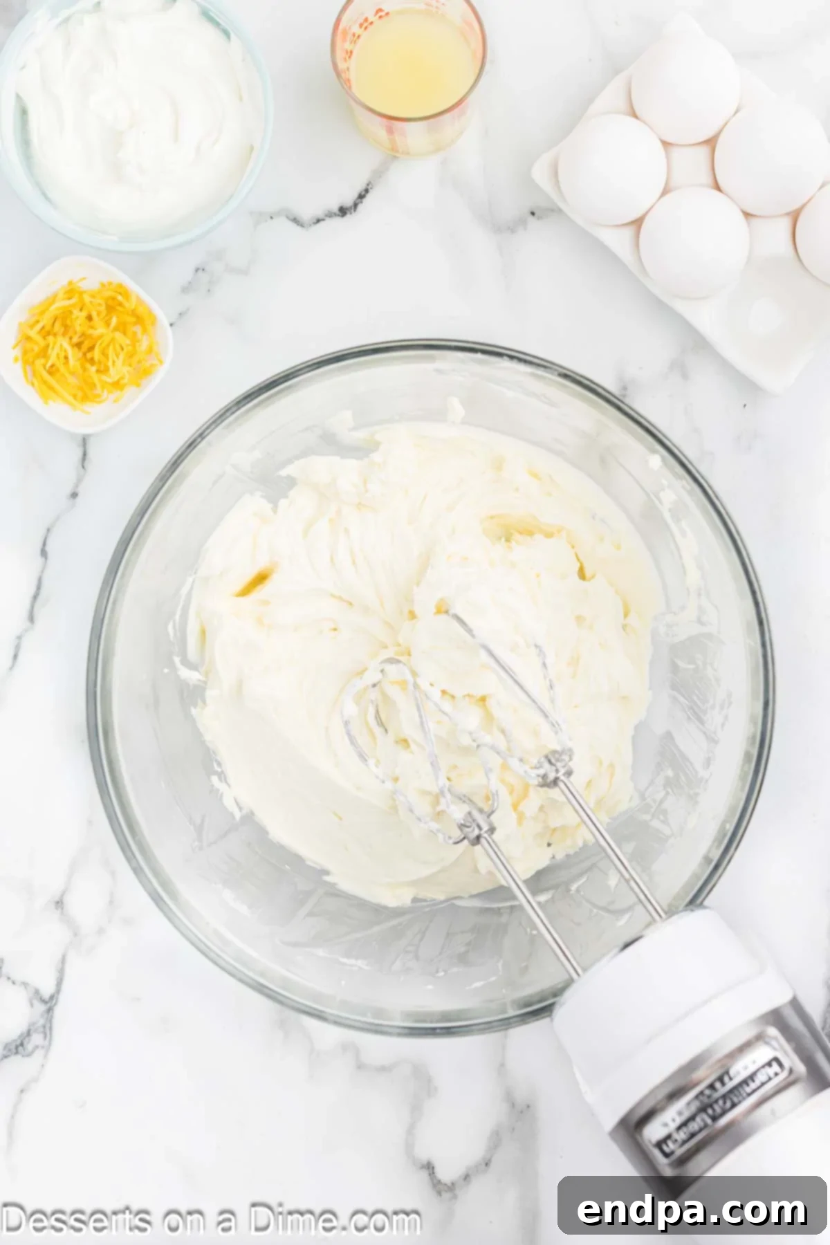 Heavenly Lemon Blueberry Cheesecake 5 A creamy, pale yellow cheesecake filling mixture is being gently beaten in a large mixing bowl with an electric mixer, showing its smooth consistency.