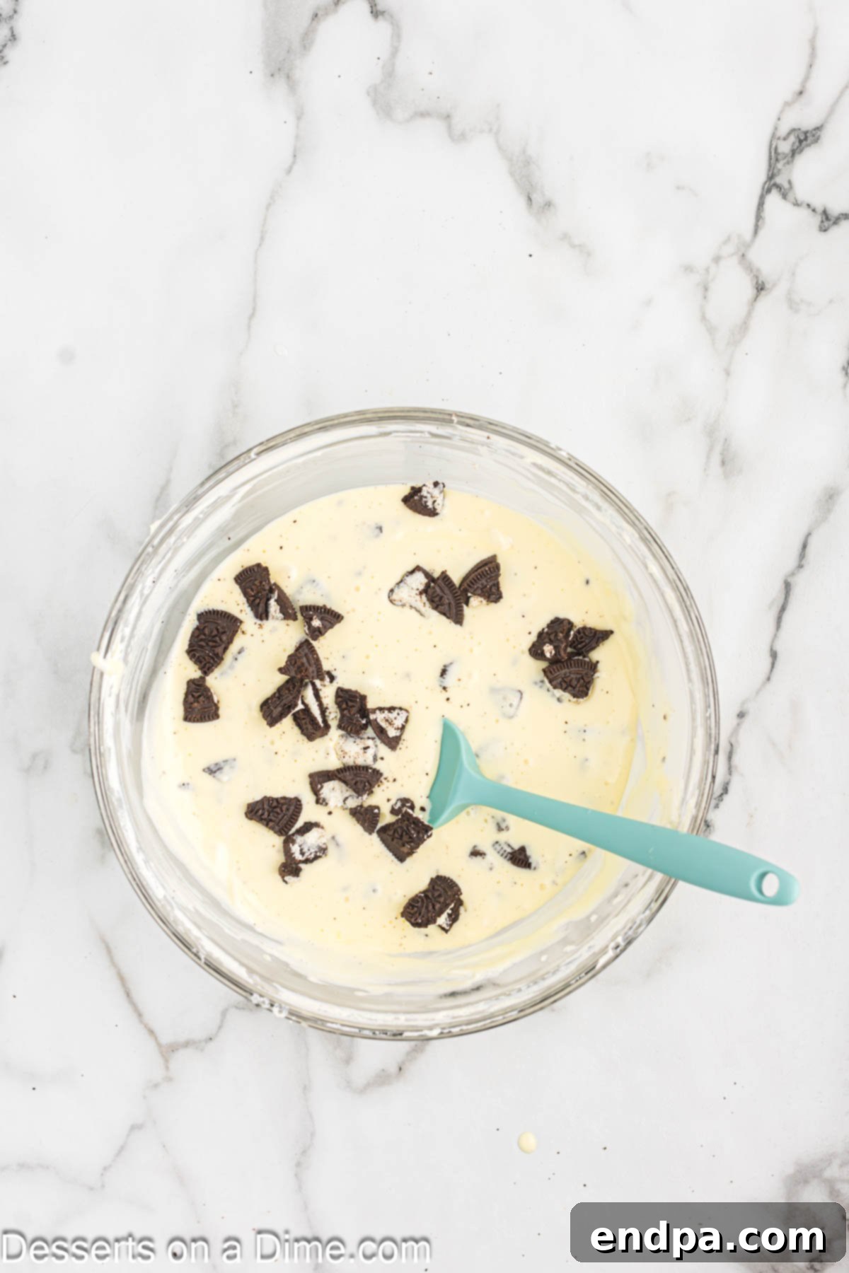 Crushed Oreo pieces being gently folded into the cheesecake mixture with a spatula by hand.