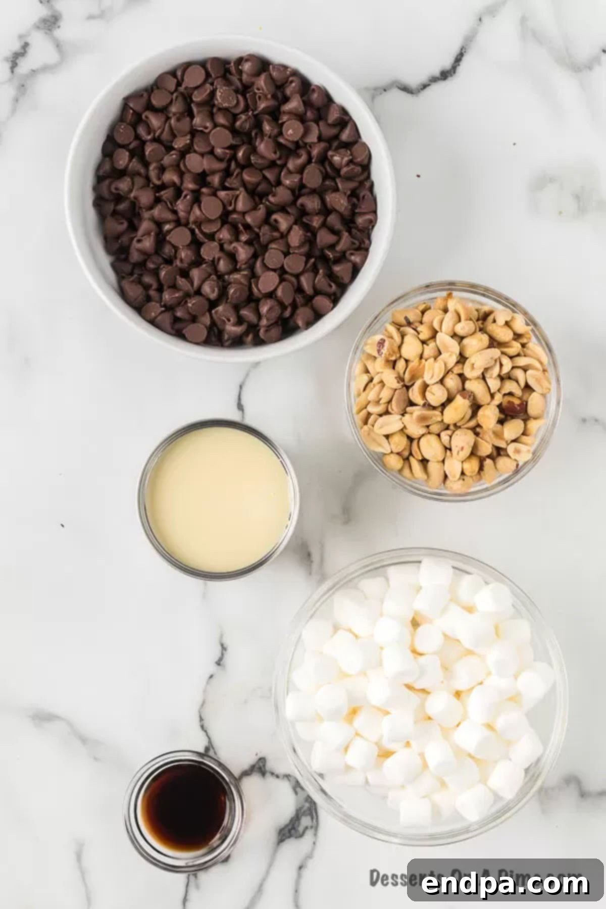 Ingredients for Rocky Road Fudge - semi sweet chocolate chips, sweetened condensed milk, vanilla, marshmallows and peanuts. All five simple ingredients neatly arranged on a counter, ready for preparation.
