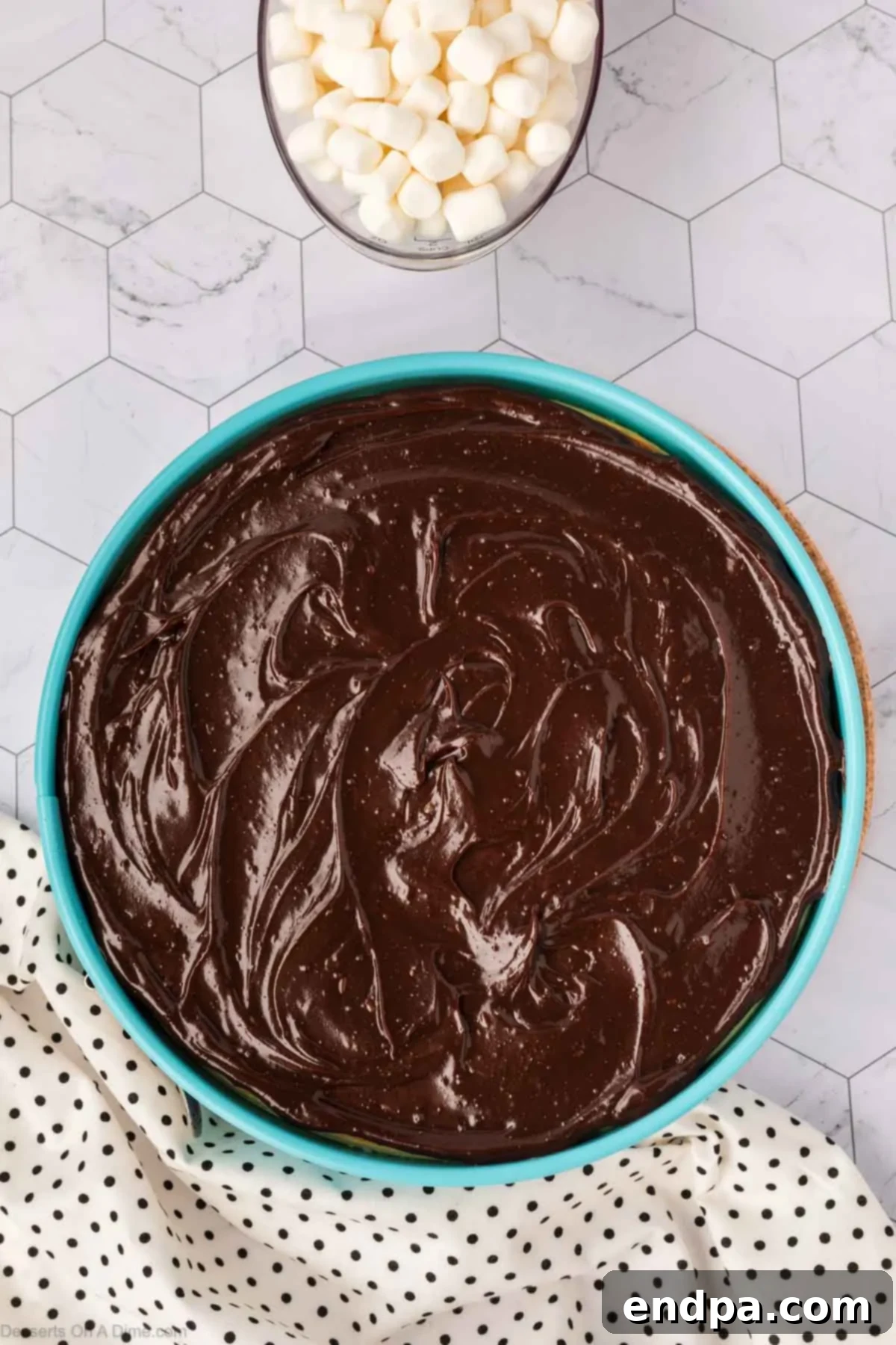 Warm hot fudge sauce being smoothly spread over the cooled cheesecake, creating a luscious chocolate layer before the marshmallow topping is added.
