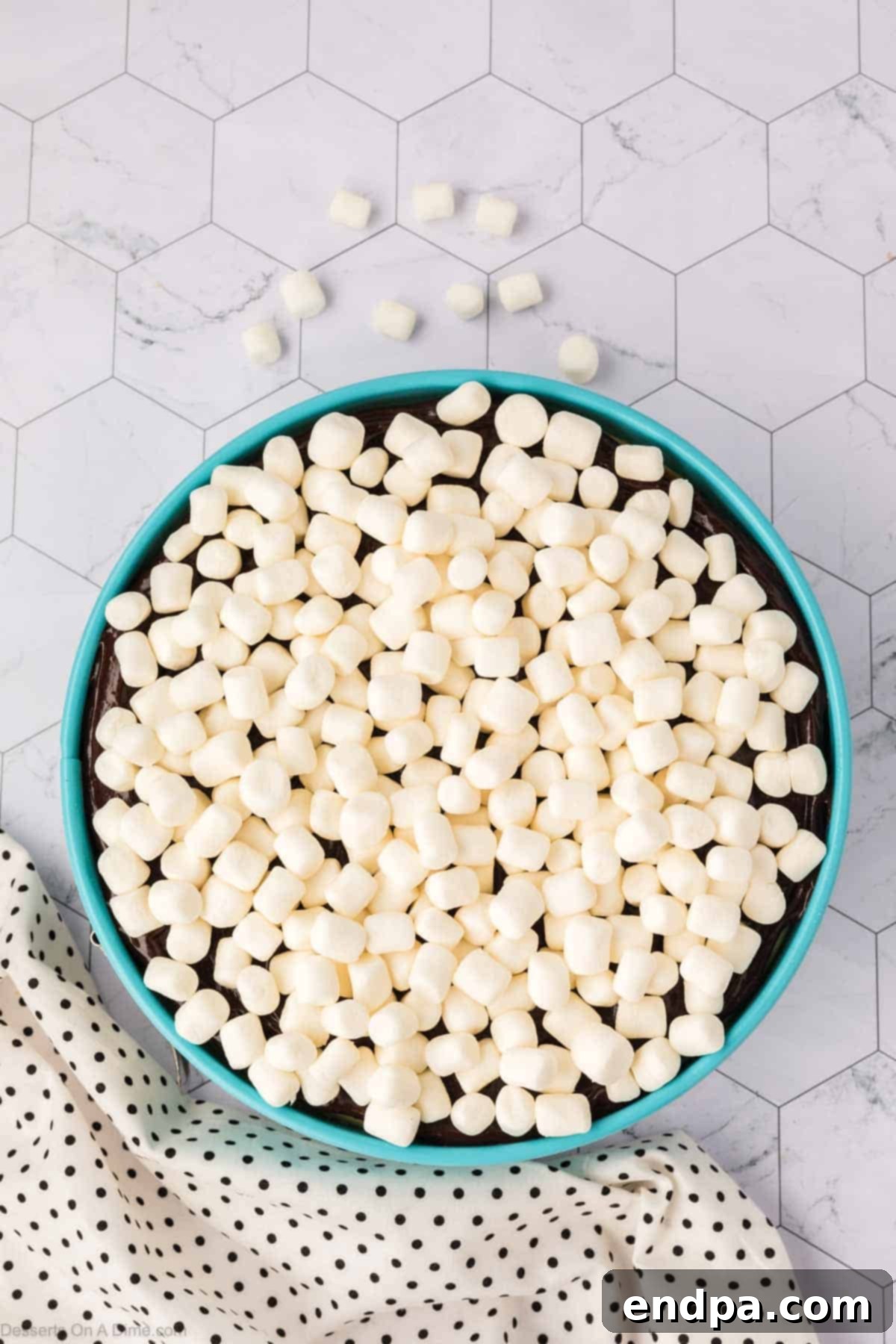 Mini marshmallows generously sprinkled over the hot fudge layer on the S'mores Cheesecake, ready for broiling to achieve a golden, toasted finish.