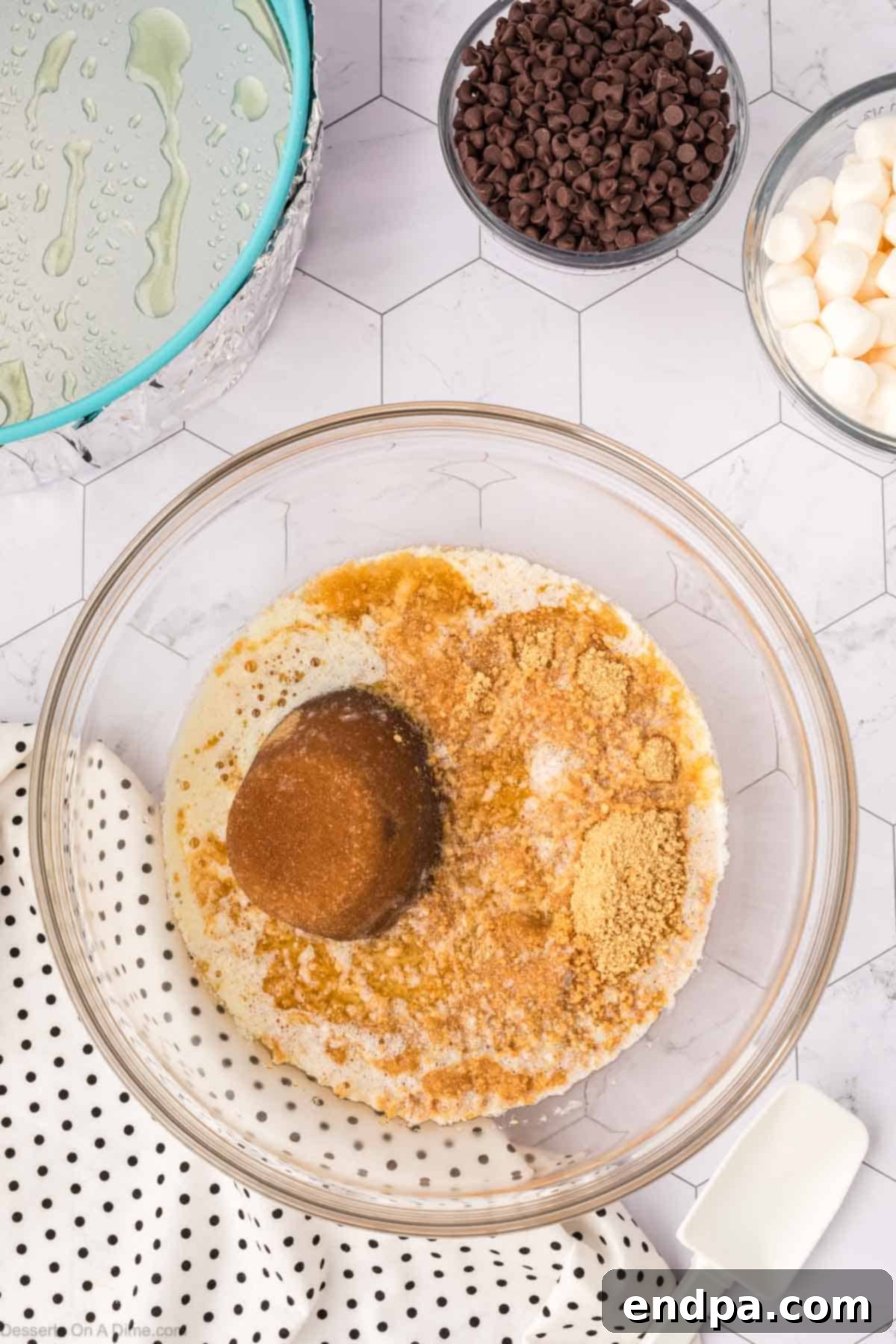 A medium mixing bowl containing graham cracker crumbs, melted butter, and brown sugar, stirred together to achieve a wet sand-like consistency for the cheesecake crust.