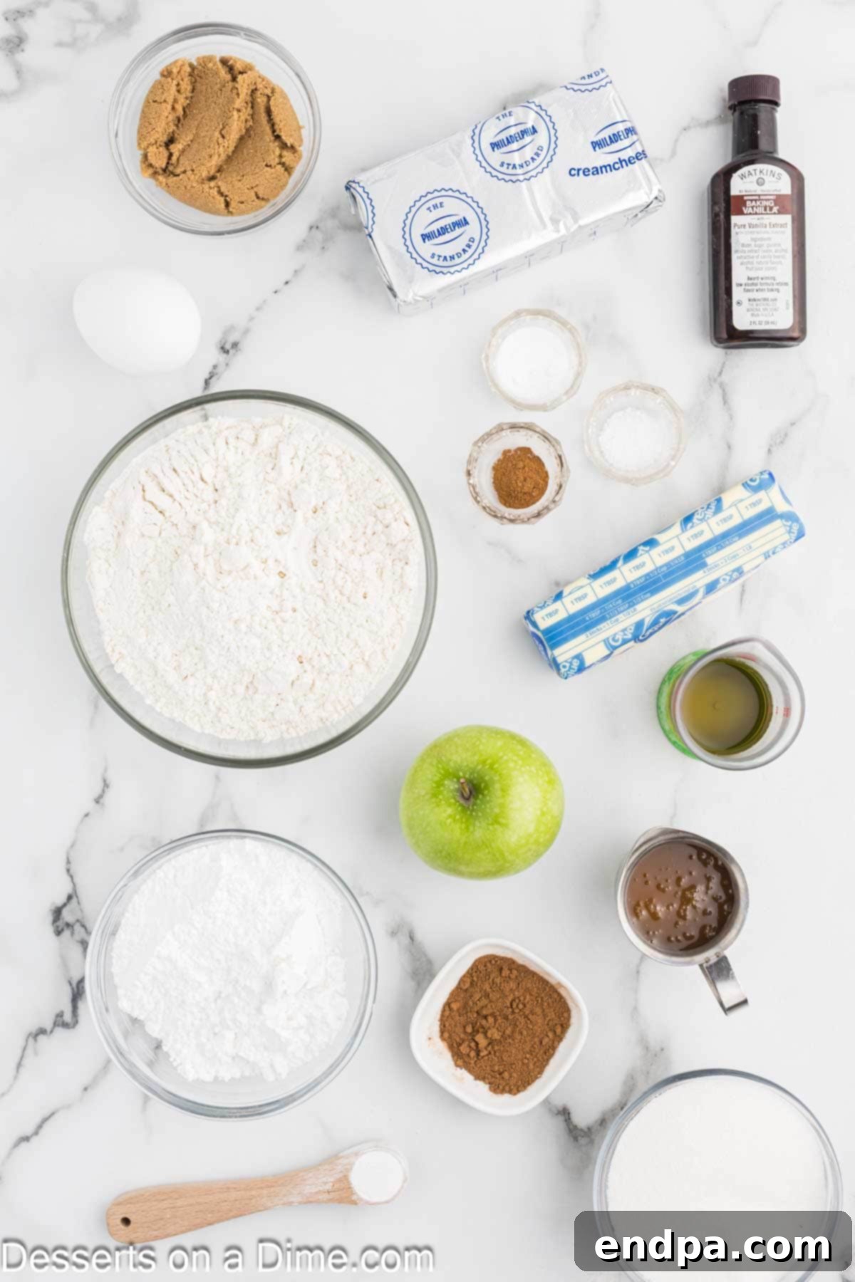 A selection of key ingredients for apple cookies, including butter, sugar, egg, apple juice, vanilla, flour, cream cheese, and caramel sauce.