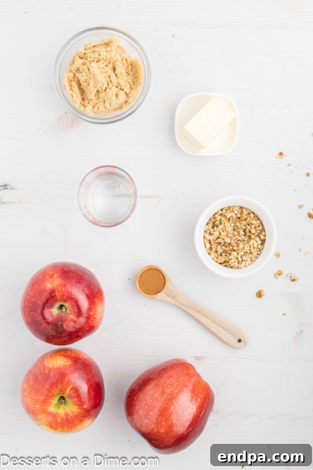 Ingredients for baked apples displayed on a wooden surface: whole red apples, a bowl of brown sugar, a container of ground cinnamon, sticks of unsalted butter, and finely chopped walnuts.