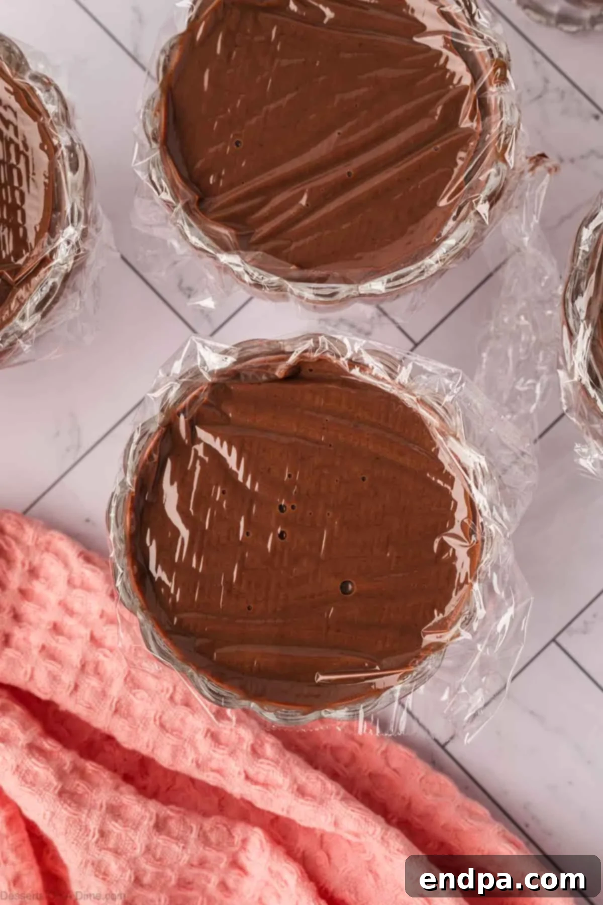 Velvety Dark Chocolate Pudding 7 Freshly cooked dark chocolate pudding is divided into six small glass cups or ramekins, each covered with plastic wrap pressed directly onto the surface to prevent a skin from forming, ready for chilling.