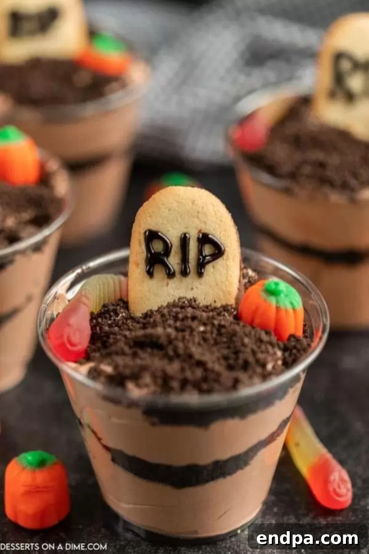 Clear plastic cup layered with chocolate pudding and Oreo cookie crumbs, topped with a Milano cookie tombstone and gummy worms.