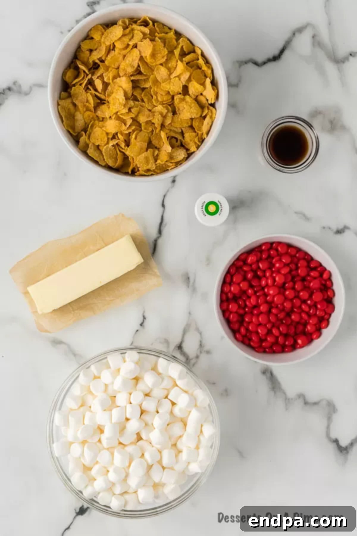 A close-up of the key ingredients laid out: cornflakes, butter, mini marshmallows, vanilla extract, green gel food coloring, and red cinnamon candies.