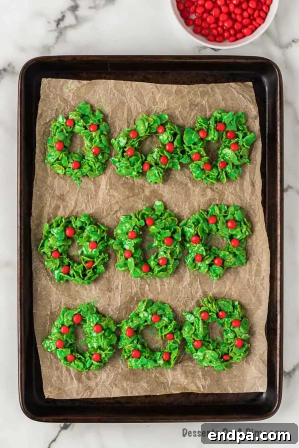 A close-up of a freshly formed green cornflake wreath, beautifully adorned with vibrant red hot candies pressed into the mixture.
