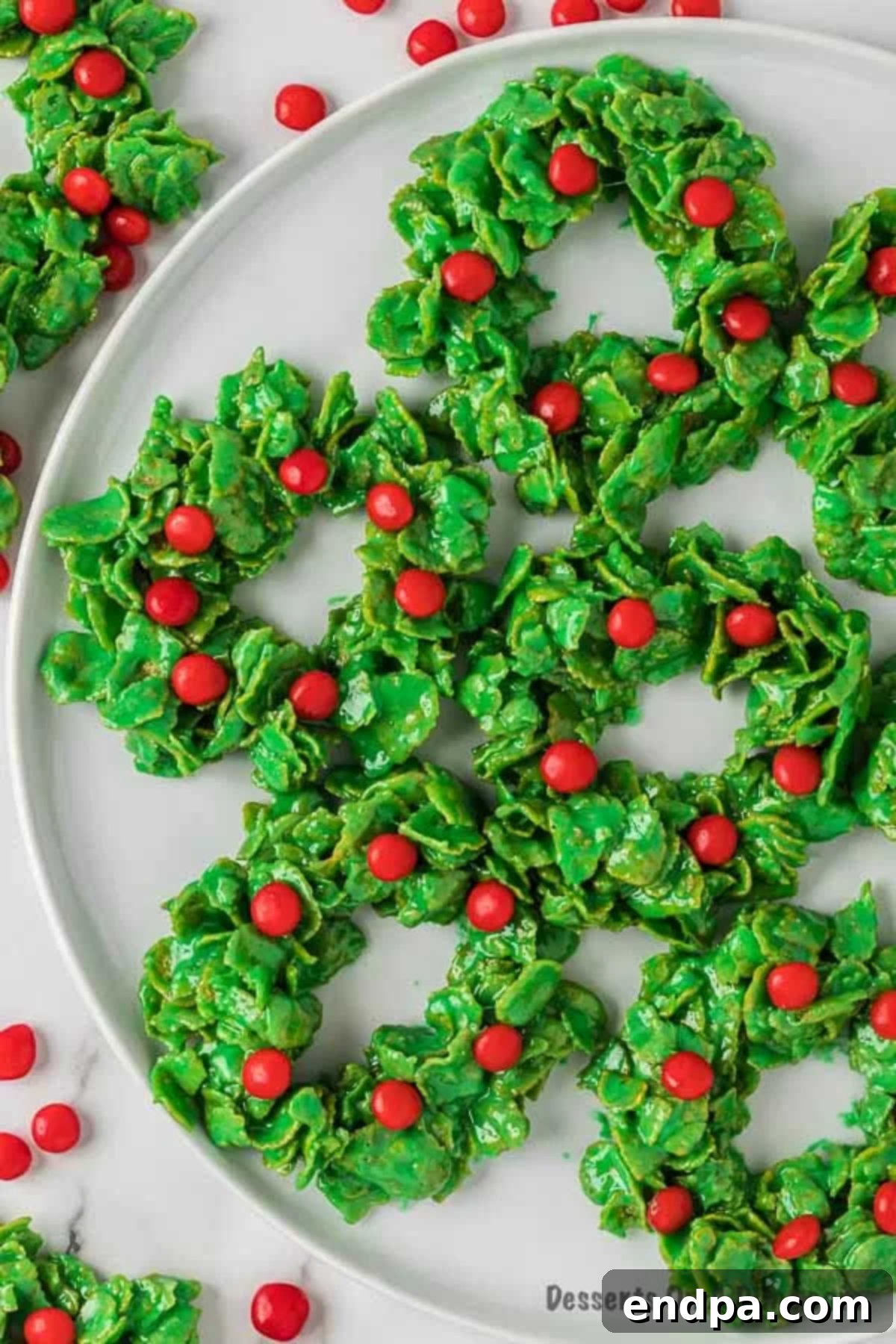 Two perfectly set Christmas wreath cookies featuring red candies, positioned in front of a platter filled with more festive cookies on a rustic wooden background.